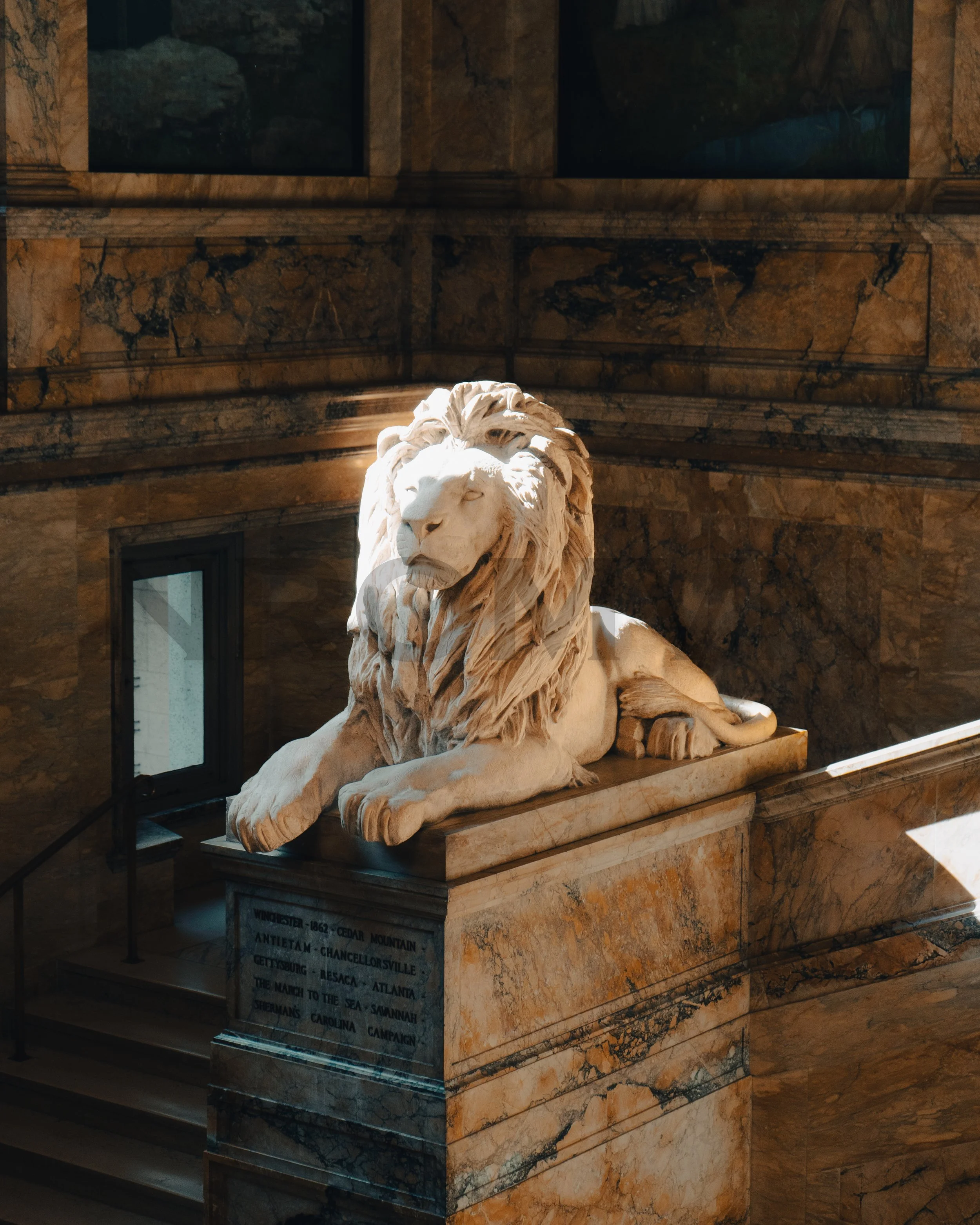 Marble statue of a reclining lion in a grand interior with brown marble walls and steps, illuminated by natural light.