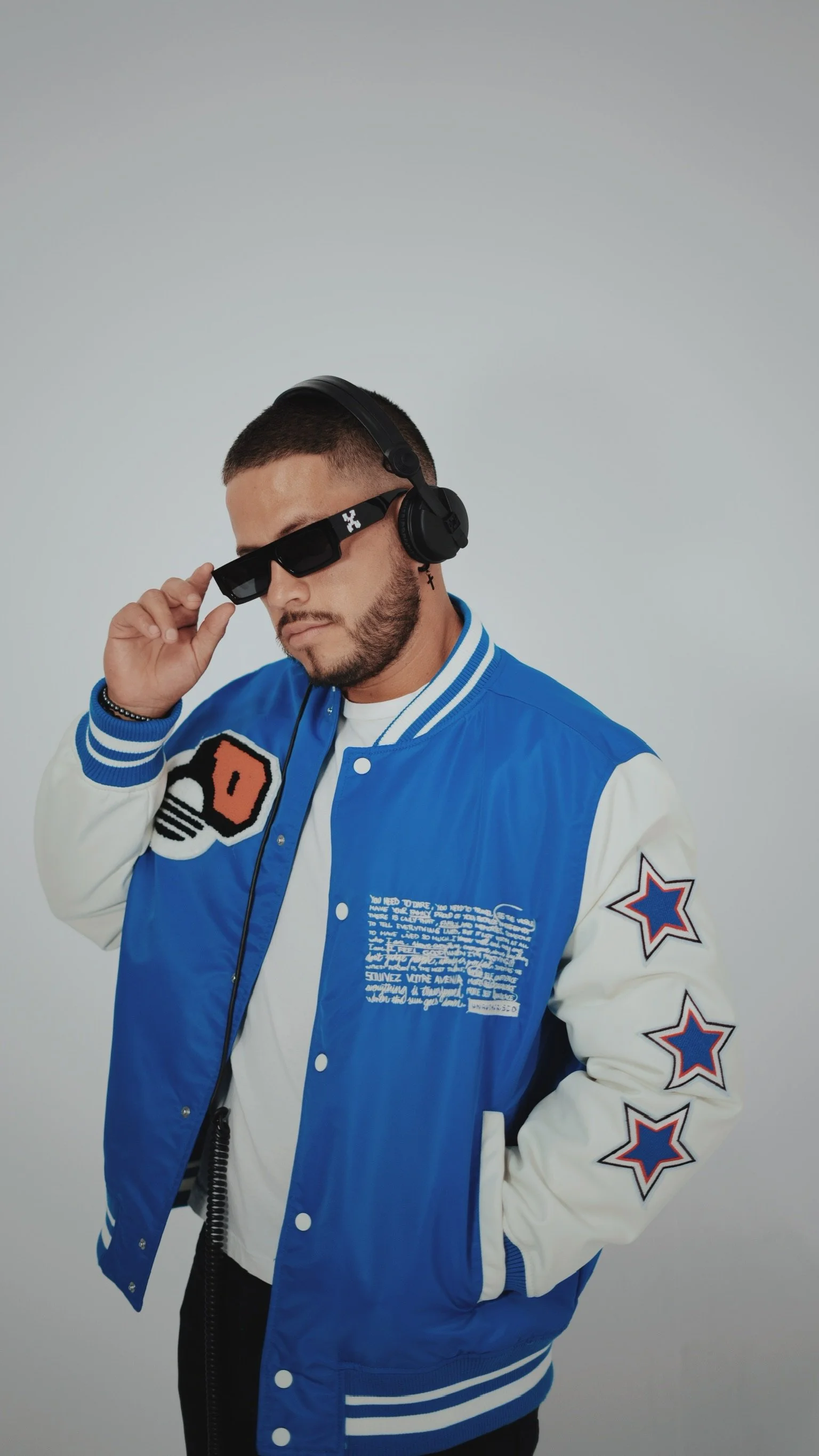 A man wearing a blue and white varsity jacket, sunglasses, and headphones, posing against a plain light gray background.