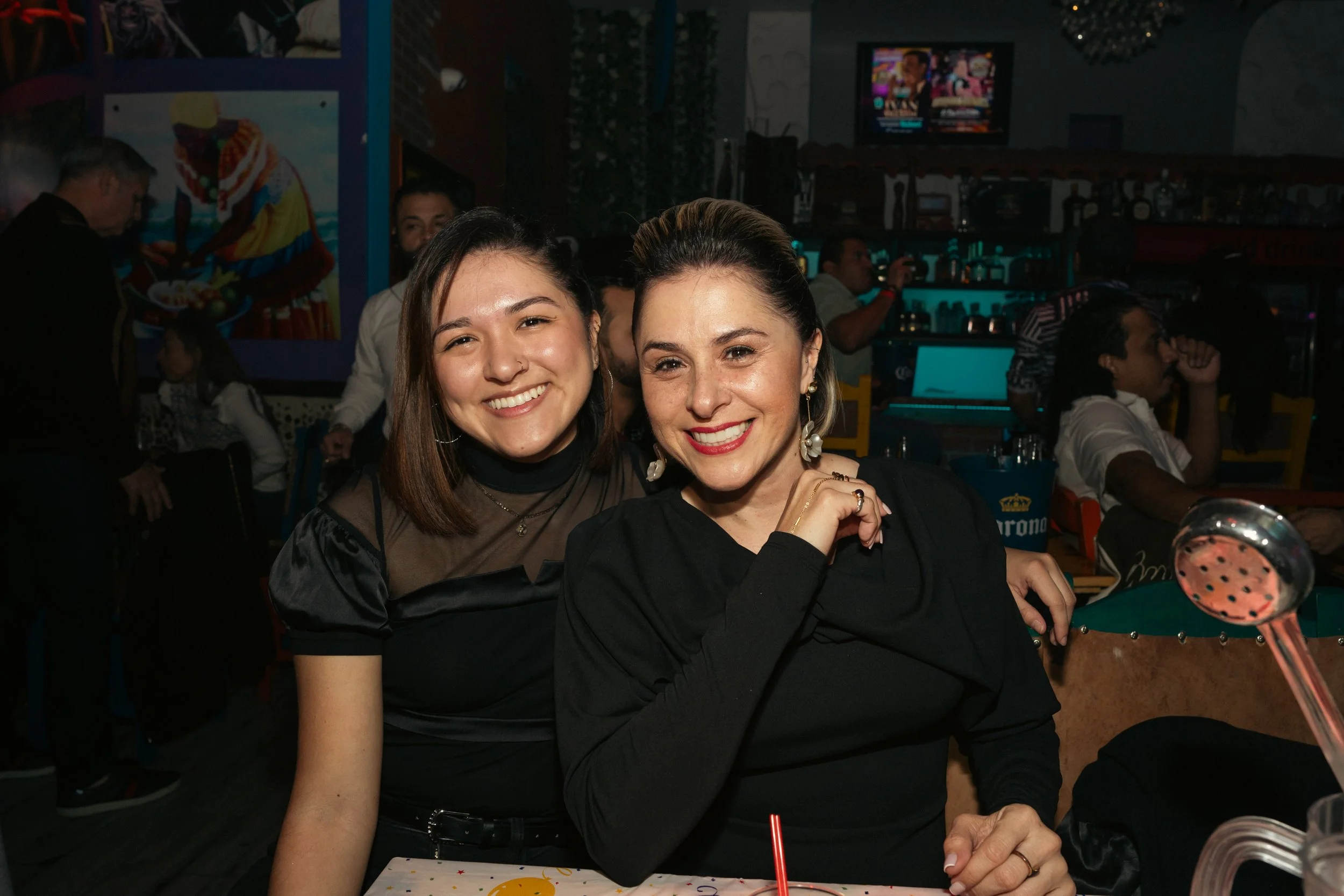 Two women smiling at a restaurant or bar, with one wearing a black top and the other wearing a brown top with black sleeves. The background shows other patrons, a television screen, and various decorations.