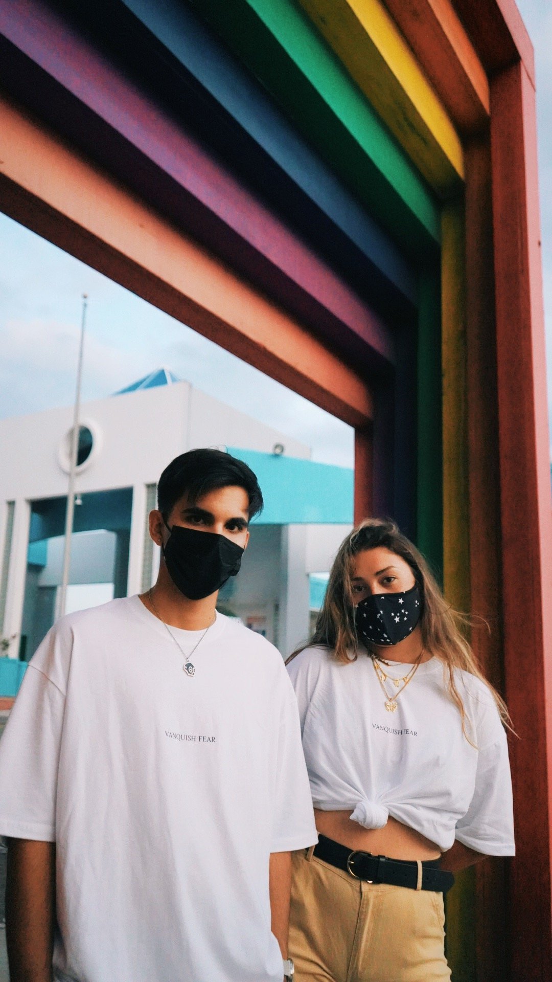 A young man and woman wearing black masks and casual clothing, standing under a colorful rainbow archway at an amusement park or fair. The woman has her shirt knotted and is wearing beige pants with a black belt, while the man is in a white T-shirt.