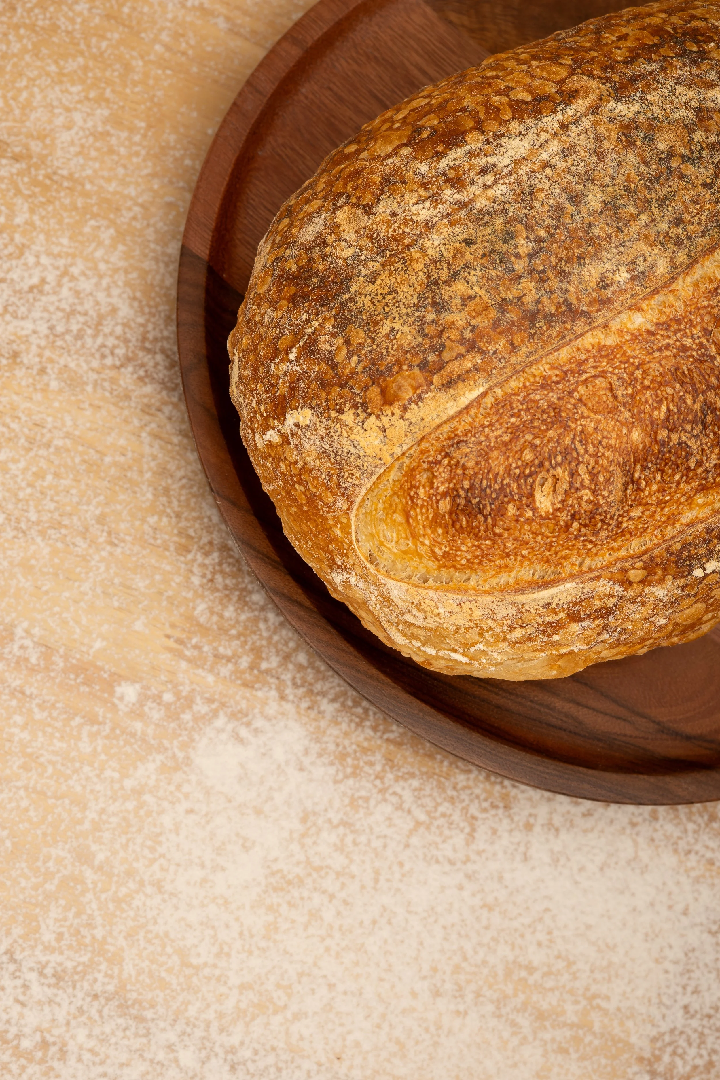 A loaf of toasted bread on a wooden plate, with a dusting of flour on the surface and surrounding area.