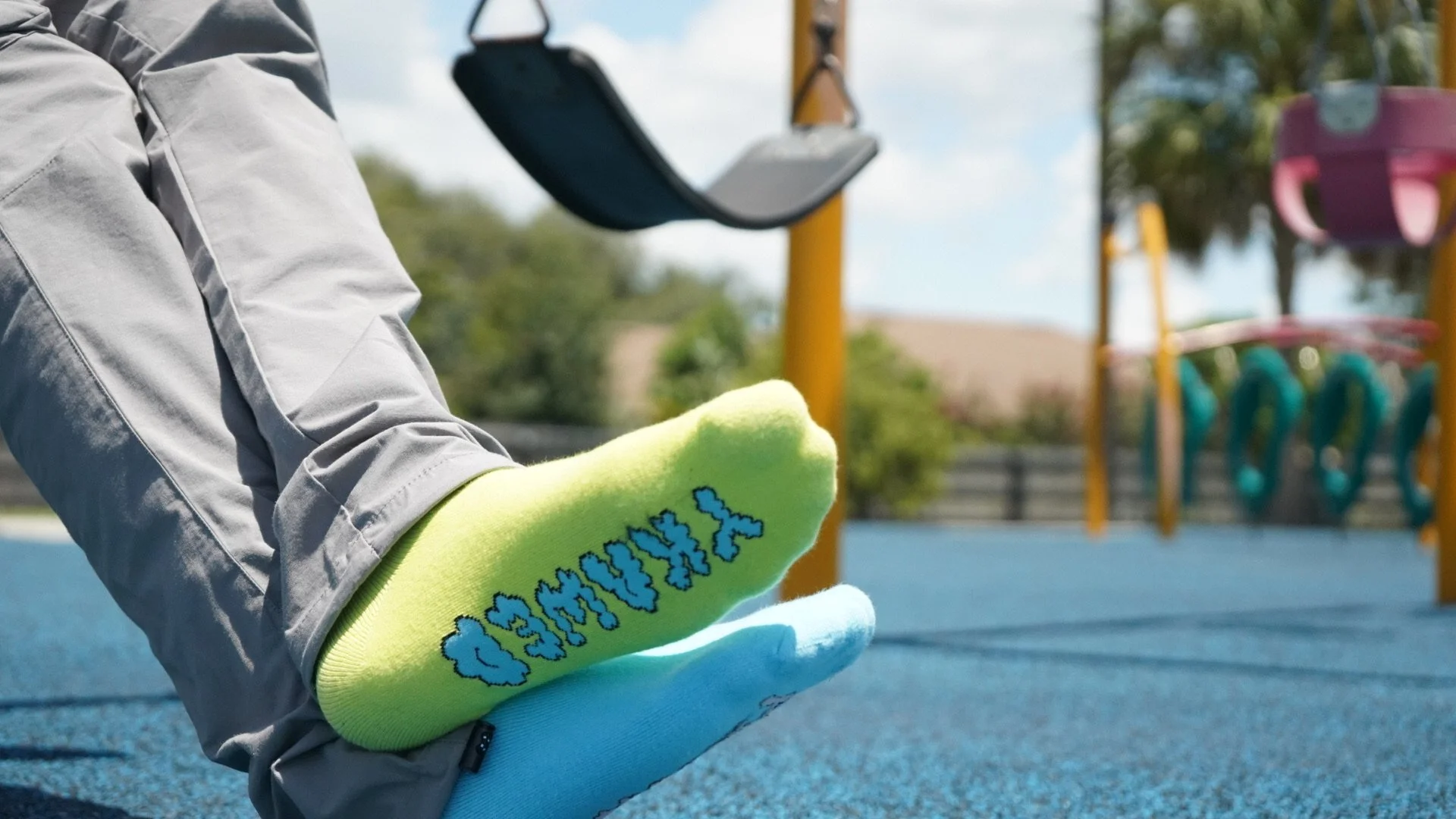 Close-up of a child's legs and feet showing neon green sock with blue lettering, and blue sock underneath, in a playground with swings in the background.