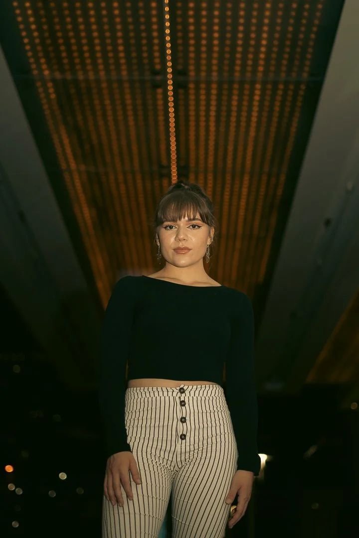 A woman standing indoors with a wooden ceiling above her, wearing a black long-sleeve top and striped high-waisted pants.