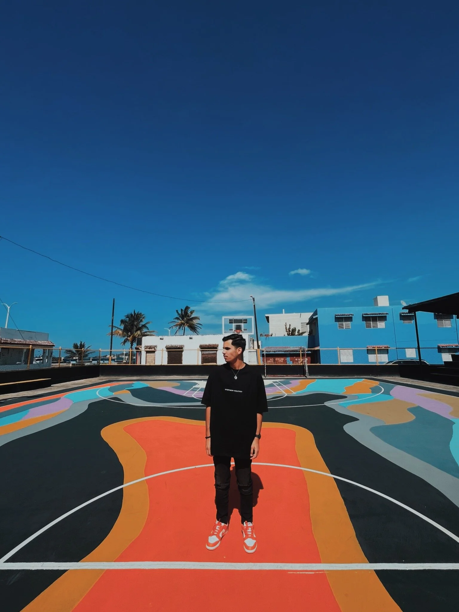A person standing on a colorful outdoor basketball court painted with abstract patterns, under a bright blue sky with a few clouds, with buildings and palm trees in the background.