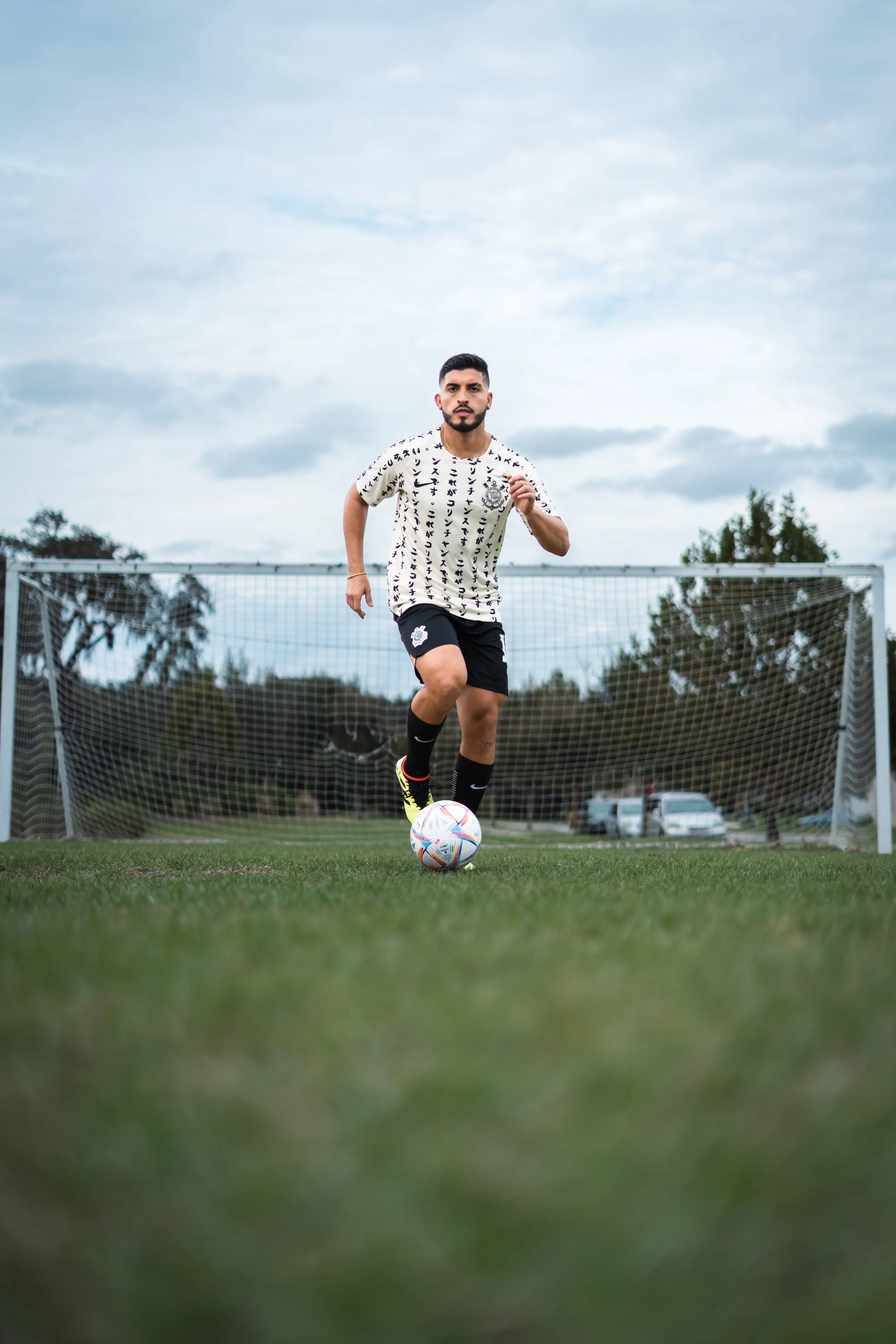 A man in a soccer uniform runs on a soccer field with a ball near a goal, with trees and cars in the background during cloudy weather.