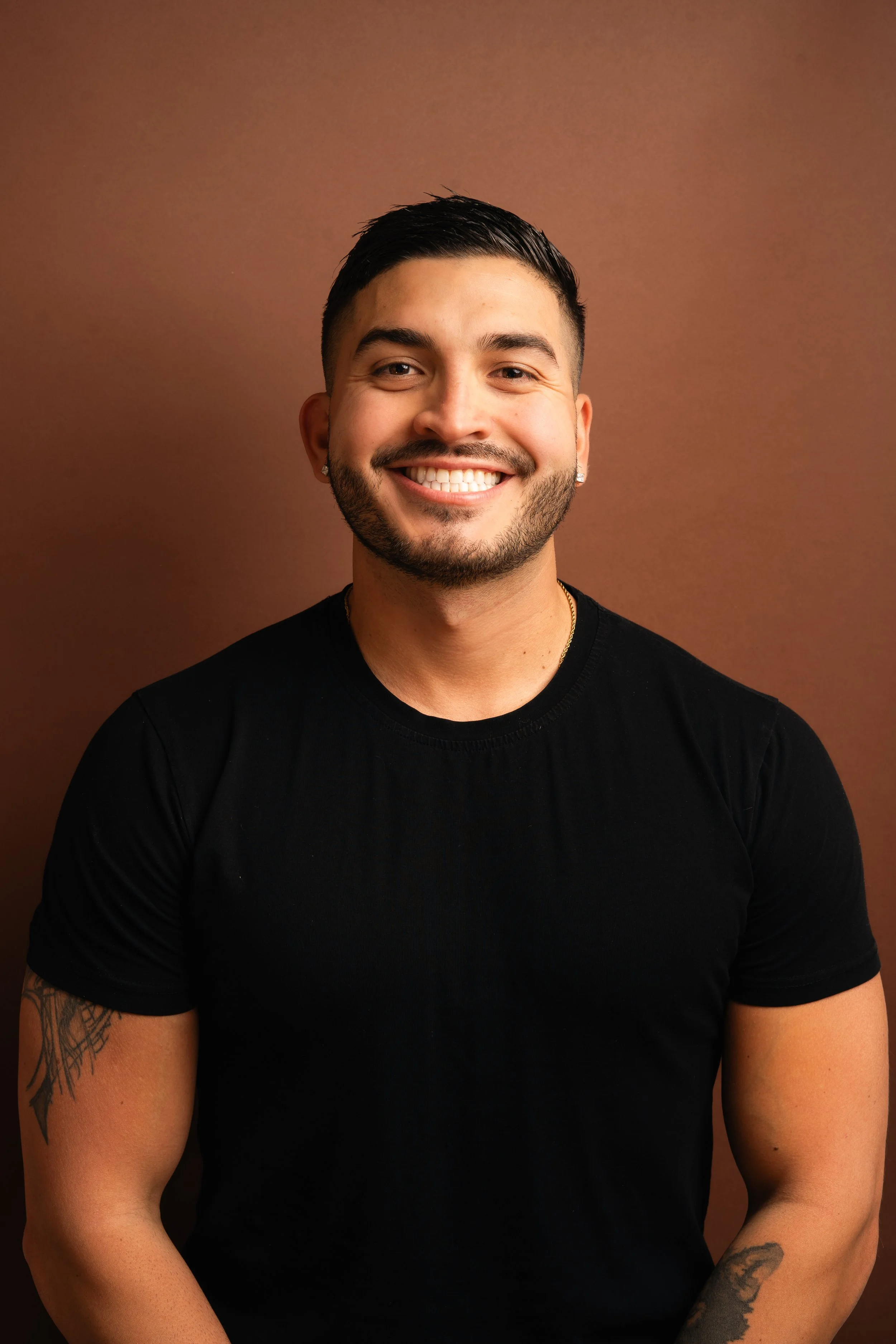 A smiling young man with short dark hair and a beard, wearing a black t-shirt, standing against a brown background.