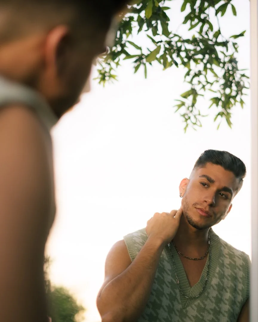 Young man with dark hair, wearing a sleeveless sweater and layered necklaces, looking into a mirror outside with trees in the background.