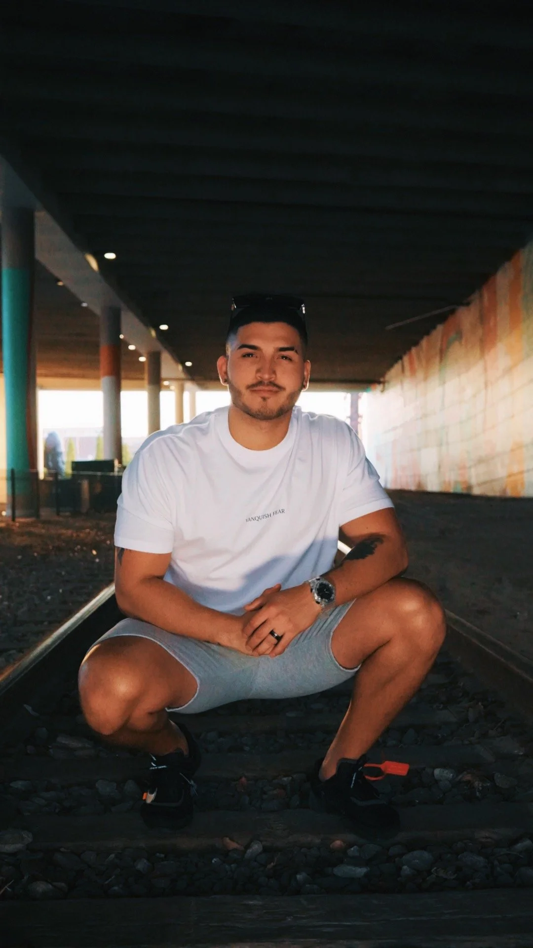 Young man with dark hair and beard squatting on railroad tracks beneath an overpass, wearing a white t-shirt, gray shorts, and black shoes, with sunglasses on his head and a watch on his wrist.