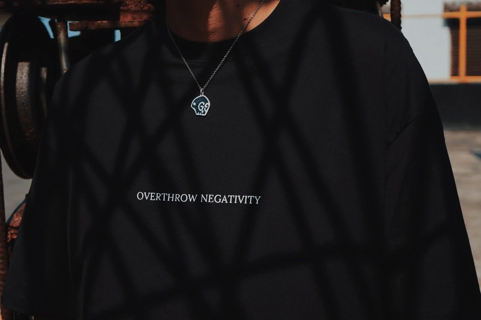 A person wearing a black t-shirt with the words 'OVERTHROW NEGATIVITY' printed on it, and a silver necklace with a skull pendant.