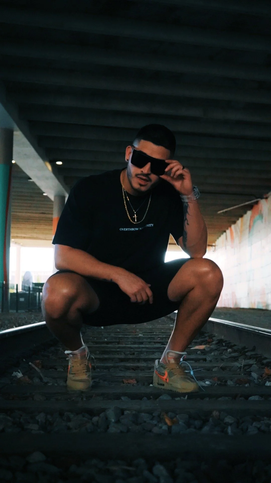A man crouching on train tracks under a bridge, wearing sunglasses, a black t-shirt, sneakers, and gold chains.