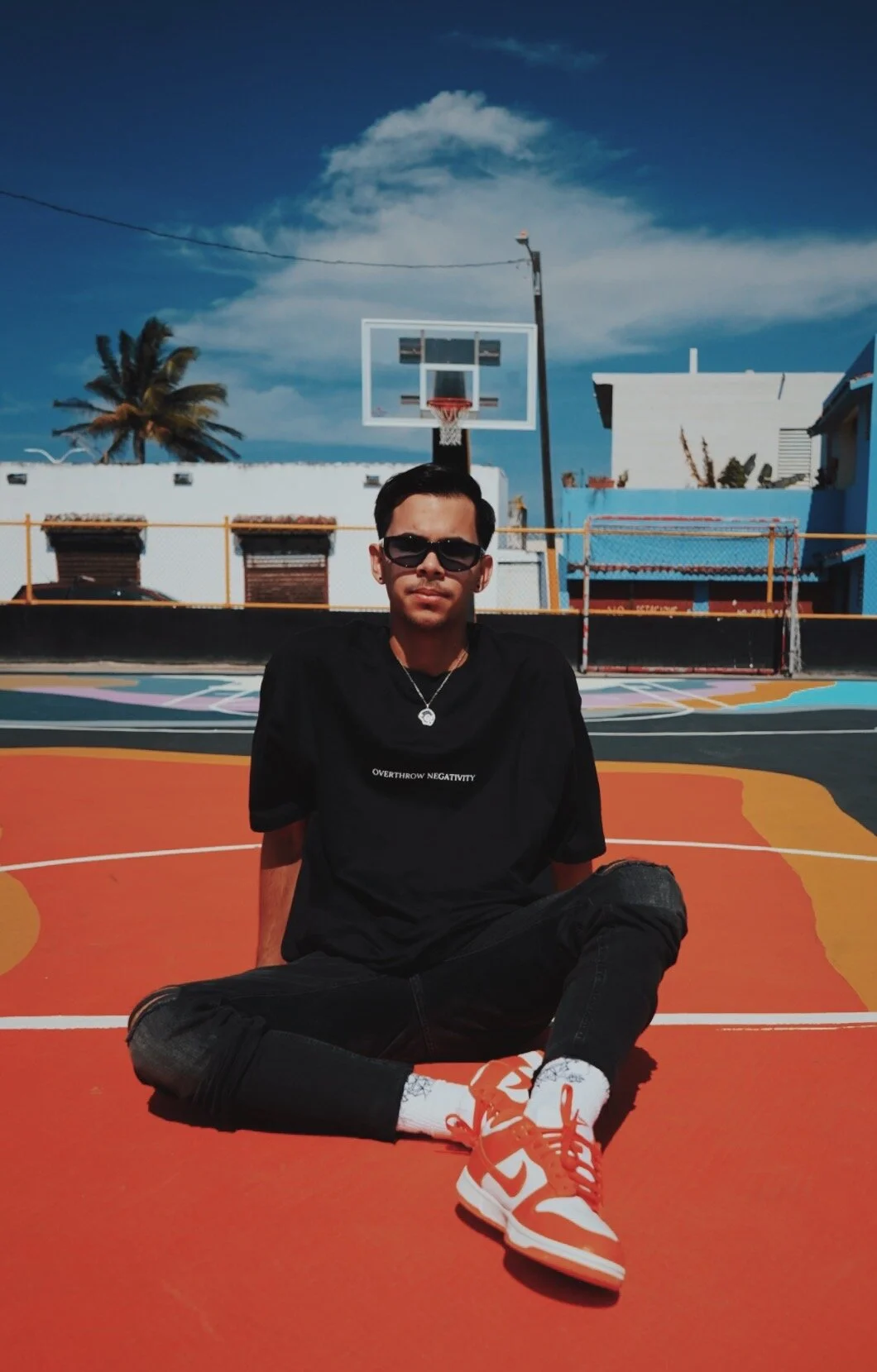 Young man sitting cross-legged on an outdoor basketball court, wearing sunglasses, black t-shirt, black jeans, and red-white sneakers. The court has a colorful surface, basketball hoop, blue sky with clouds, and palm trees in the background.