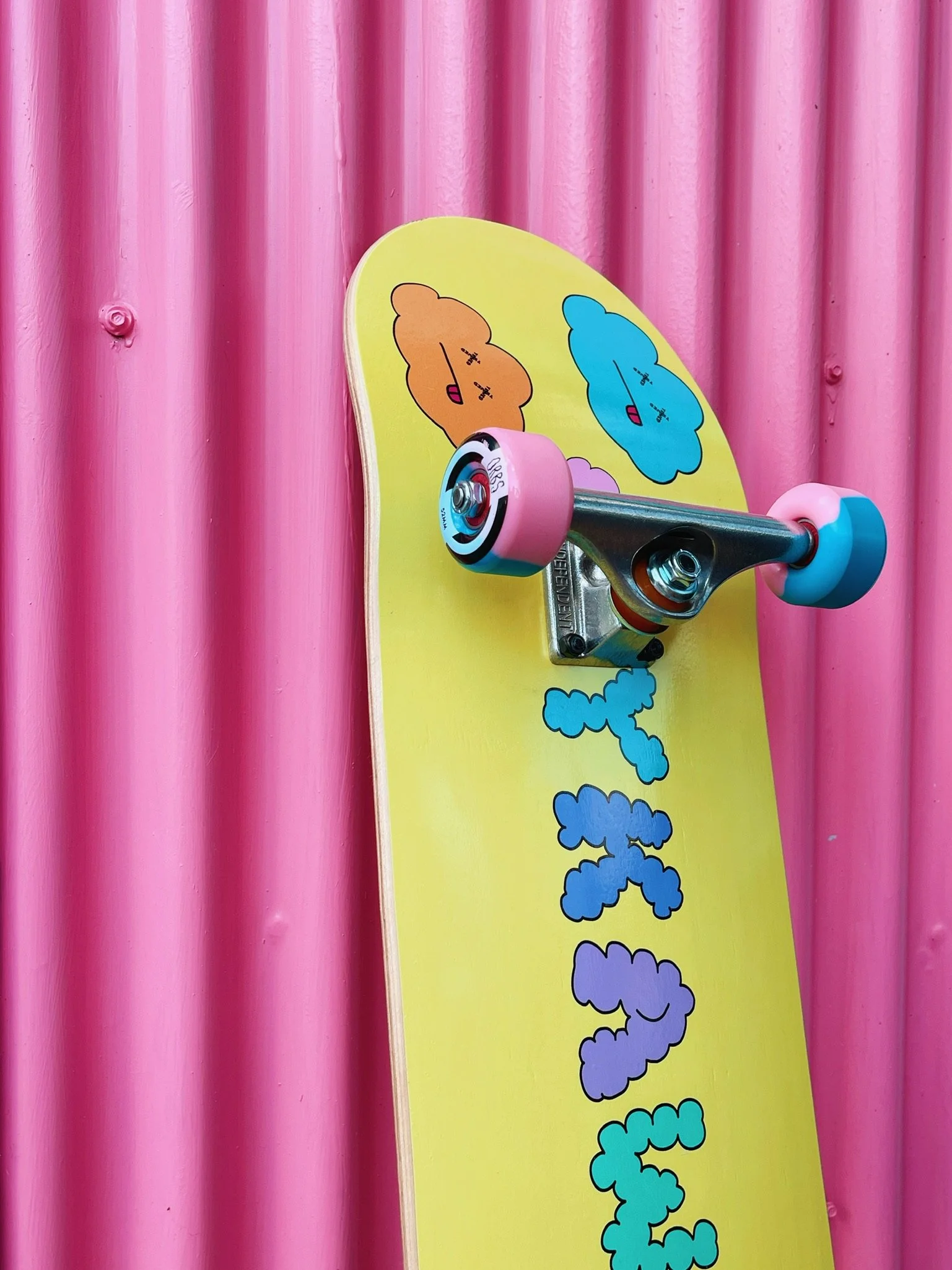 A yellow skateboard with colorful cloud characters and the words 'Cloudy' on it, mounted on a pink corrugated metal wall.
