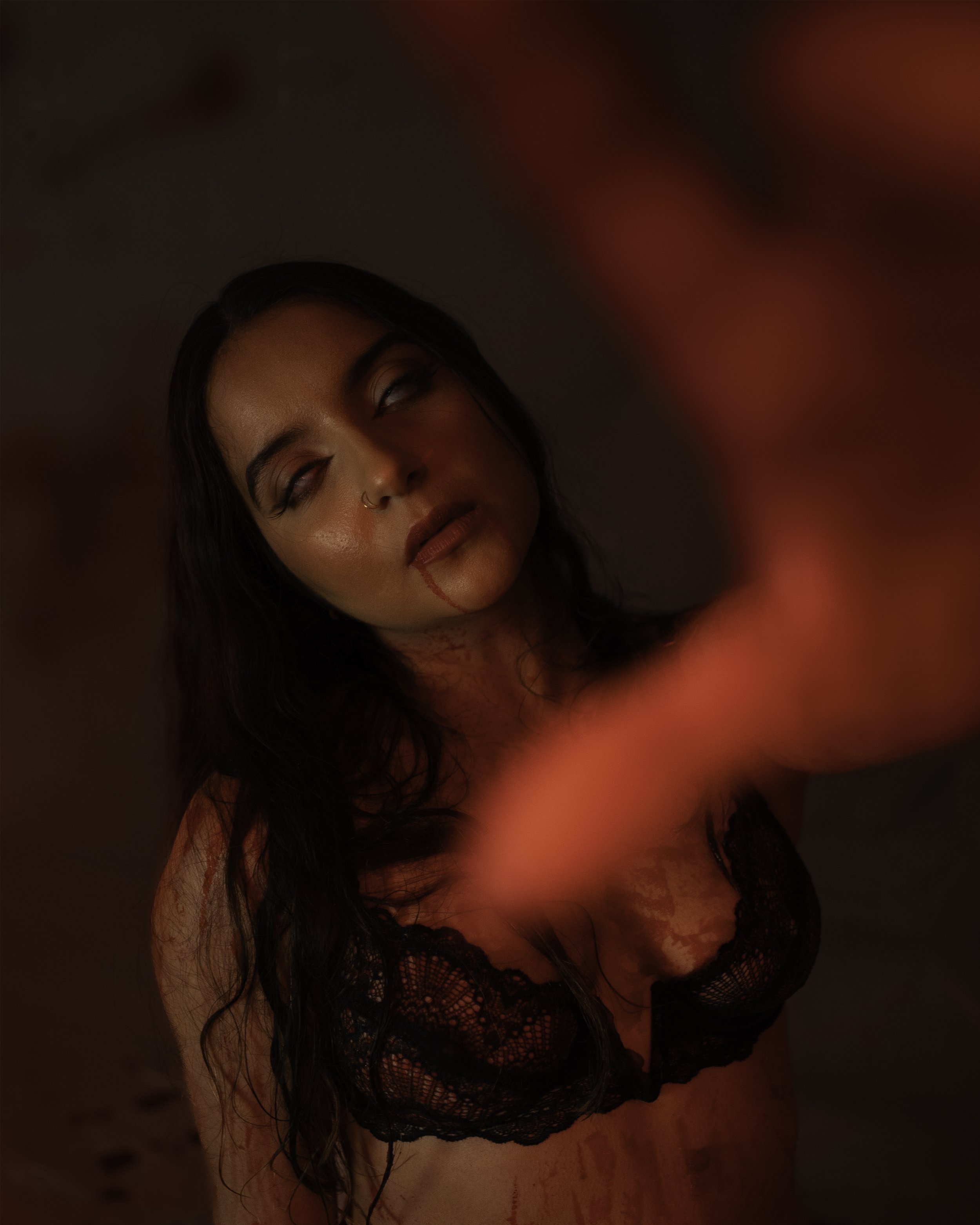 A woman with long dark hair, wearing a black lace top, looks upward with her eyes half-closed. She has a nose ring and red lipstick, and there is red lighting on her face and body, with a blurred red background.