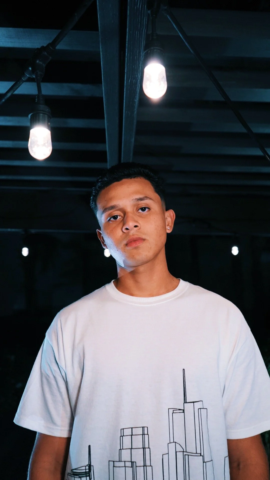 A young man with short black hair and earrings posing under a ceiling with hanging light bulbs in a dark setting.
