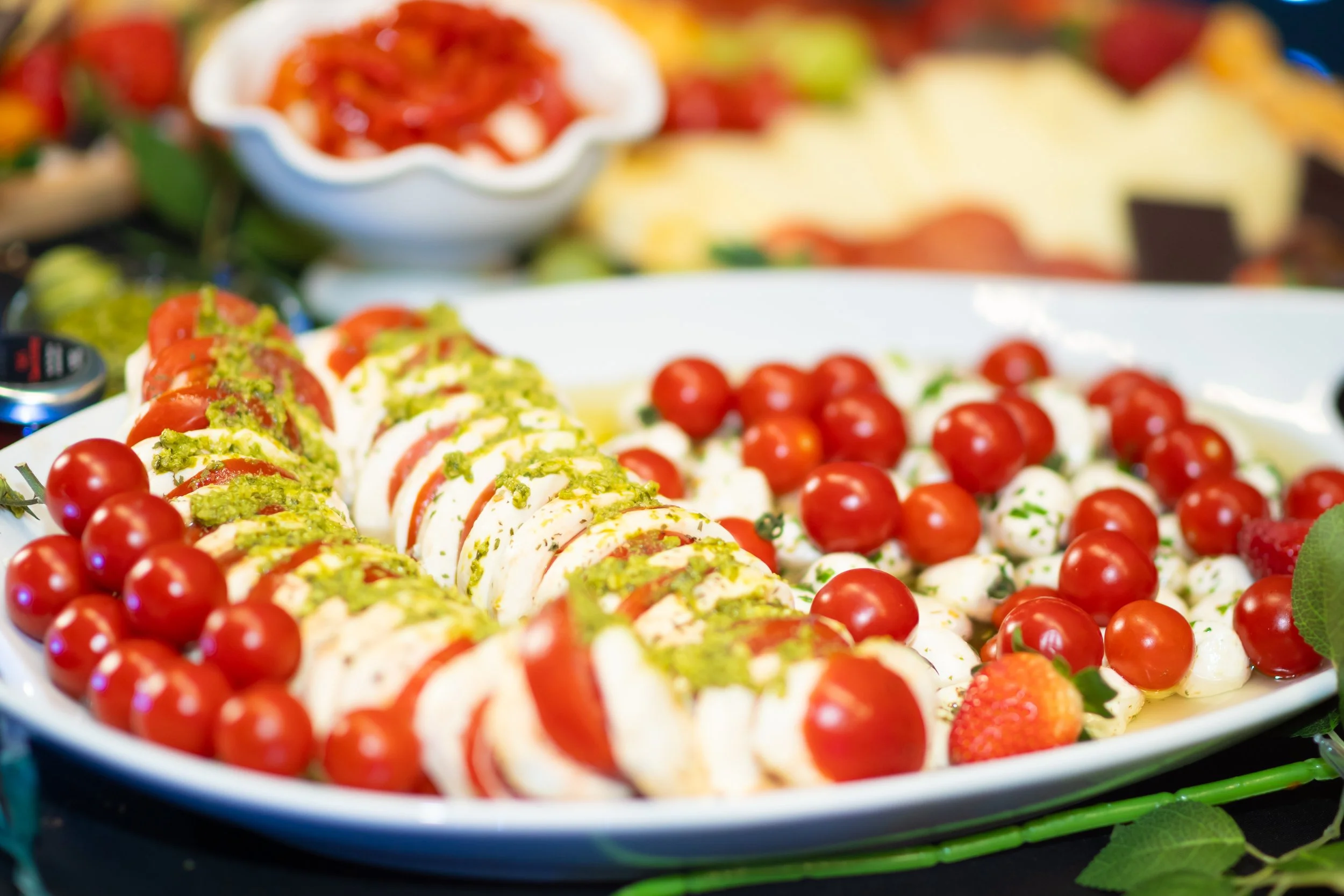 A white platter with sliced mozzarella and cherry tomatoes topped with chopped green herbs, alongside whole cherry tomatoes, in a colorful food display.