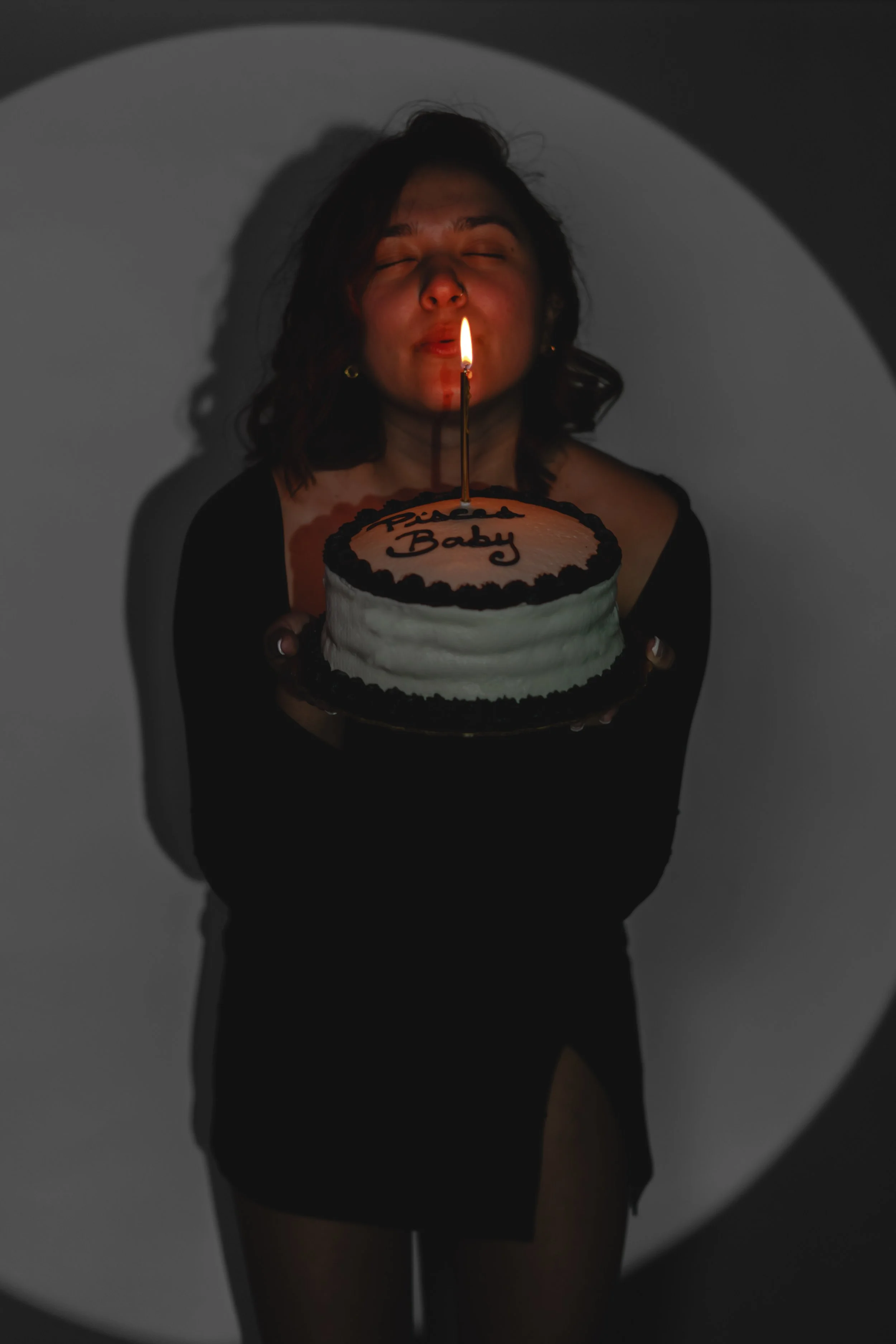A woman with short curly hair holds a birthday cake with a lit candle, smiling with her eyes closed. The cake has white frosting and the words 'Happy Baby' written on top.