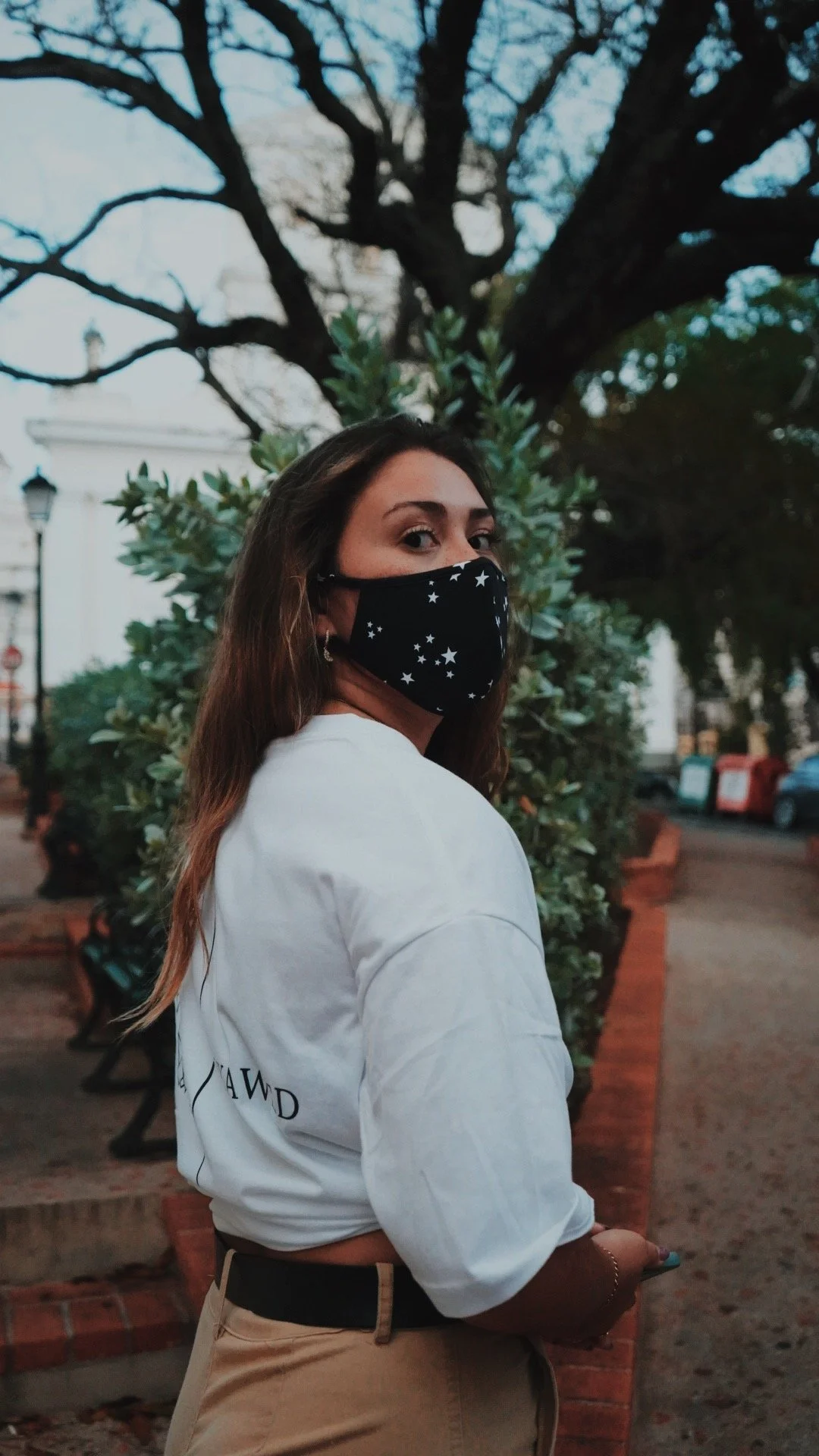 A woman with brown hair wearing a black face mask with white stars, a white long-sleeve shirt, and beige pants, standing outdoors near a brick sidewalk, with trees and parked cars in the background.