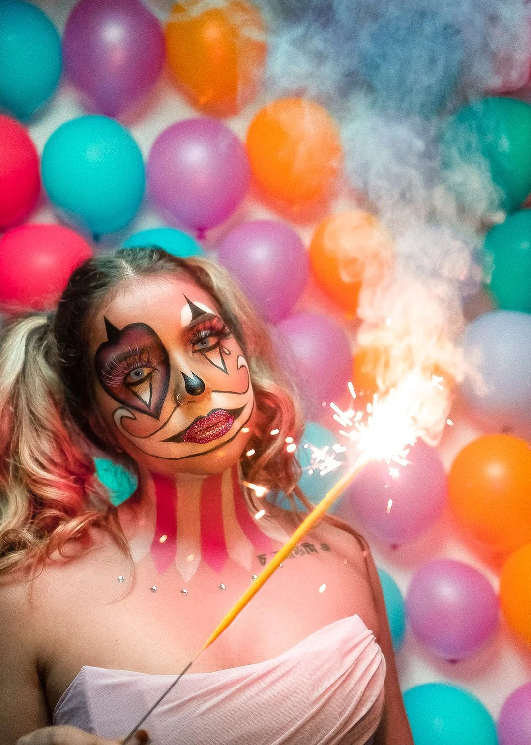 A woman with face makeup resembling a sugar skull, holding a lit sparkler, surrounded by colorful balloons and smoke.