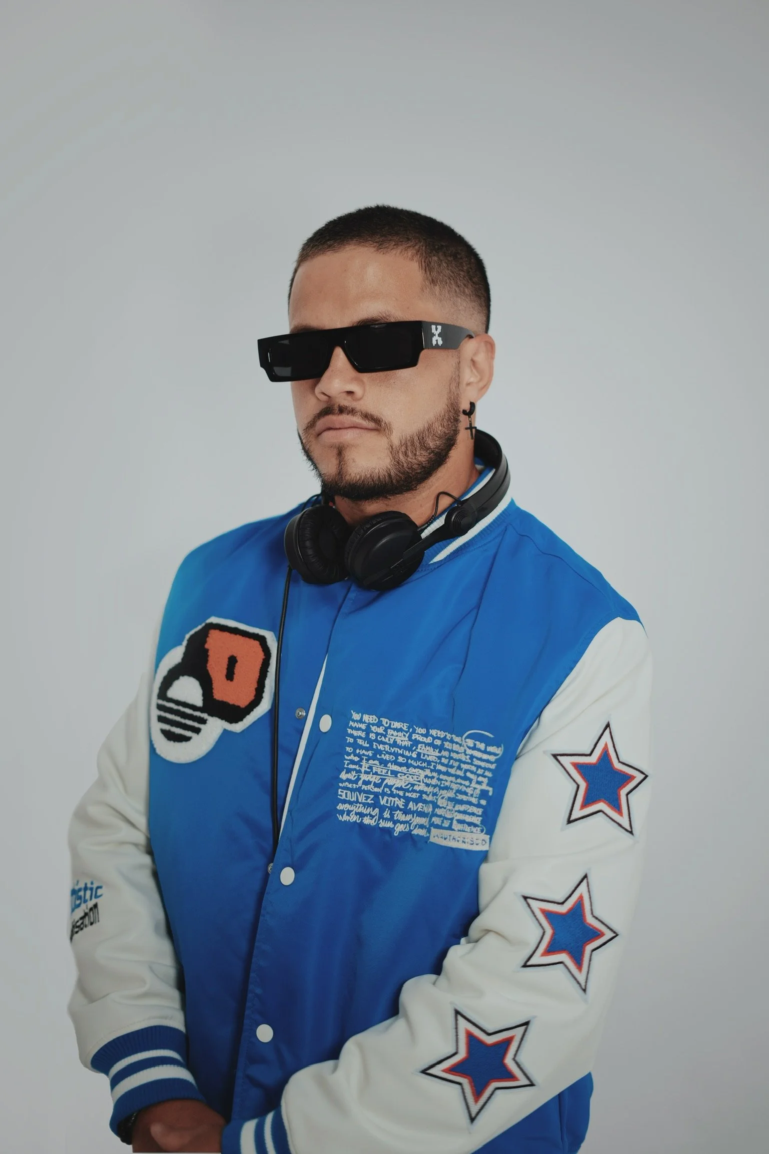 A young man with a beard wearing black sunglasses, headphones around his neck, and a blue and white varsity jacket with patches and text, posing against a plain gray background.