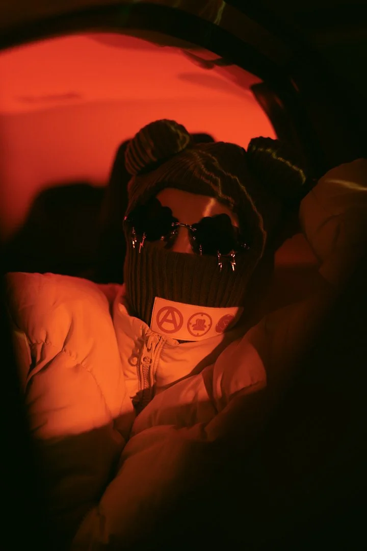 Person wearing sunglasses, a beanie, and a jacket, with a face covering that has symbols on it, inside a car with red lighting.