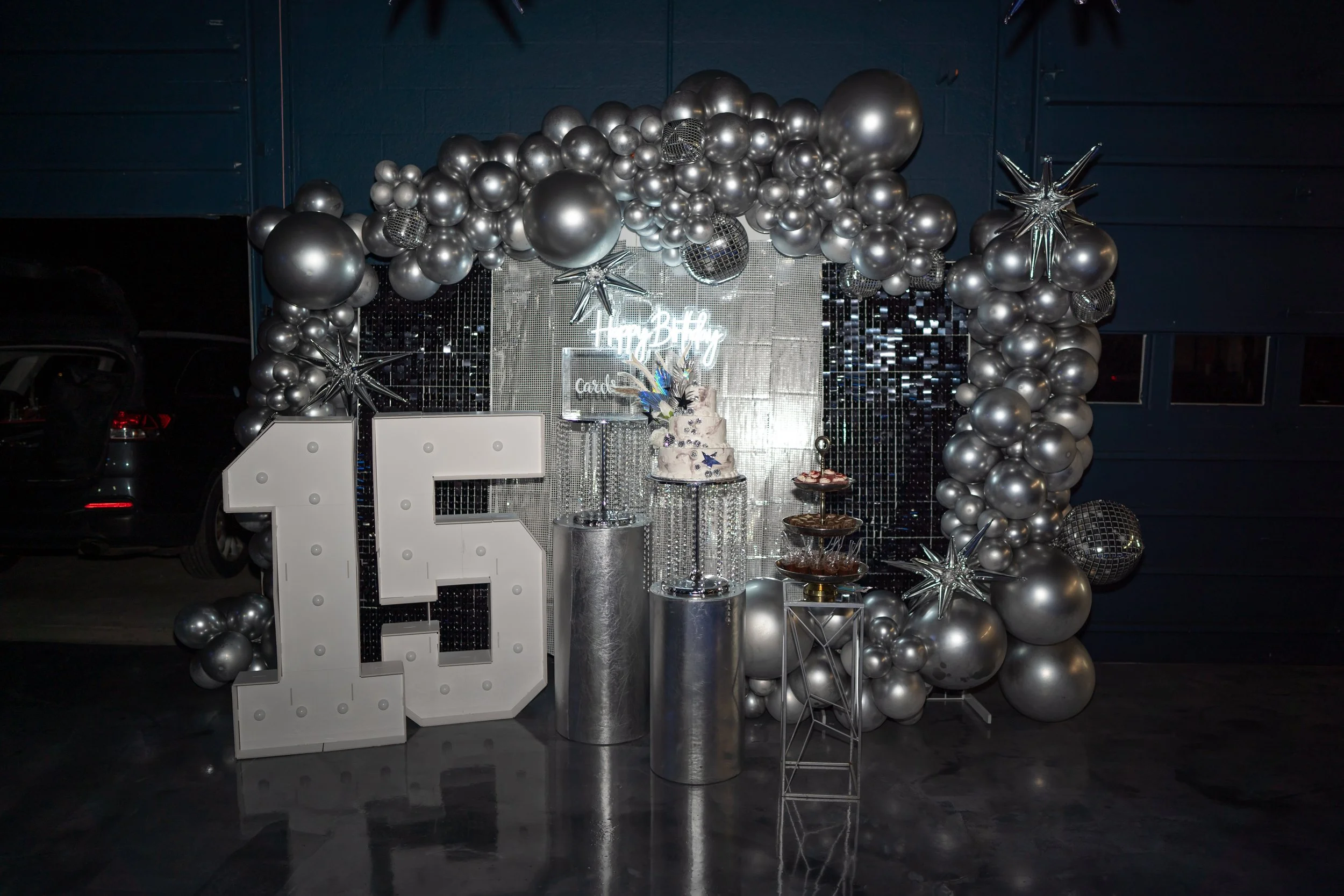 Decorative 16th birthday party setup with silver balloons, a large numbered 16, a cake on a stand, and a metallic backdrop with a 'Happy Birthday' sign.