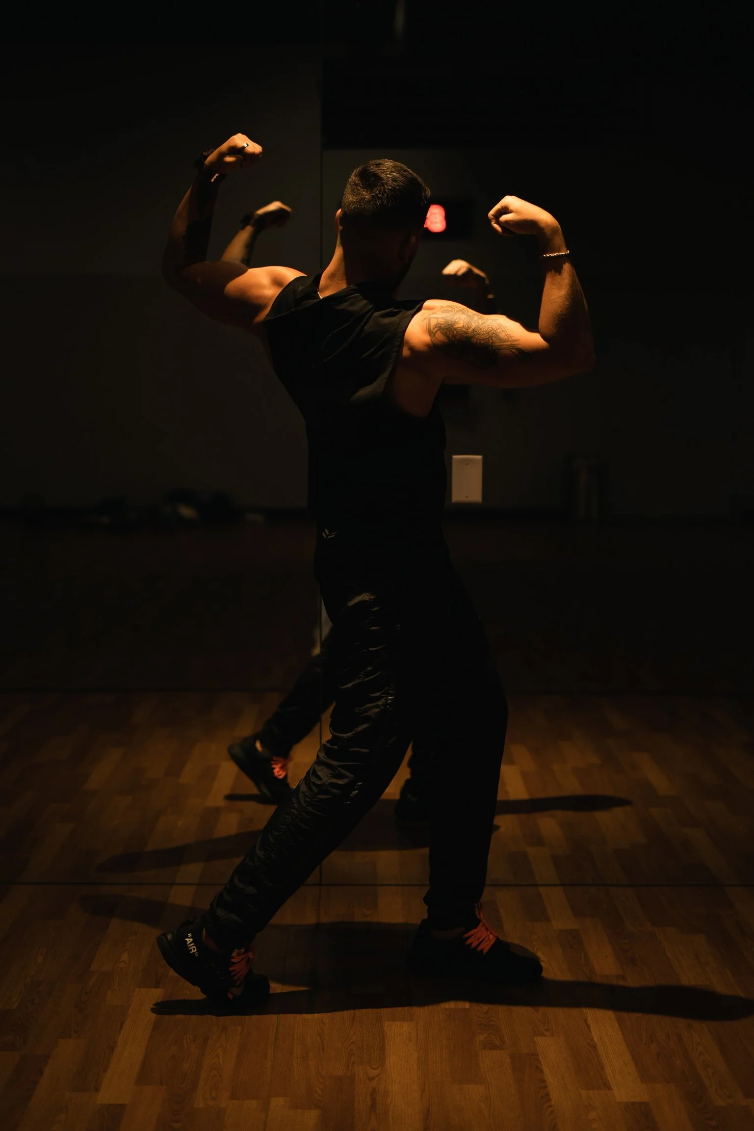 A muscular man flexing his biceps in a dimly lit room with a wooden floor.