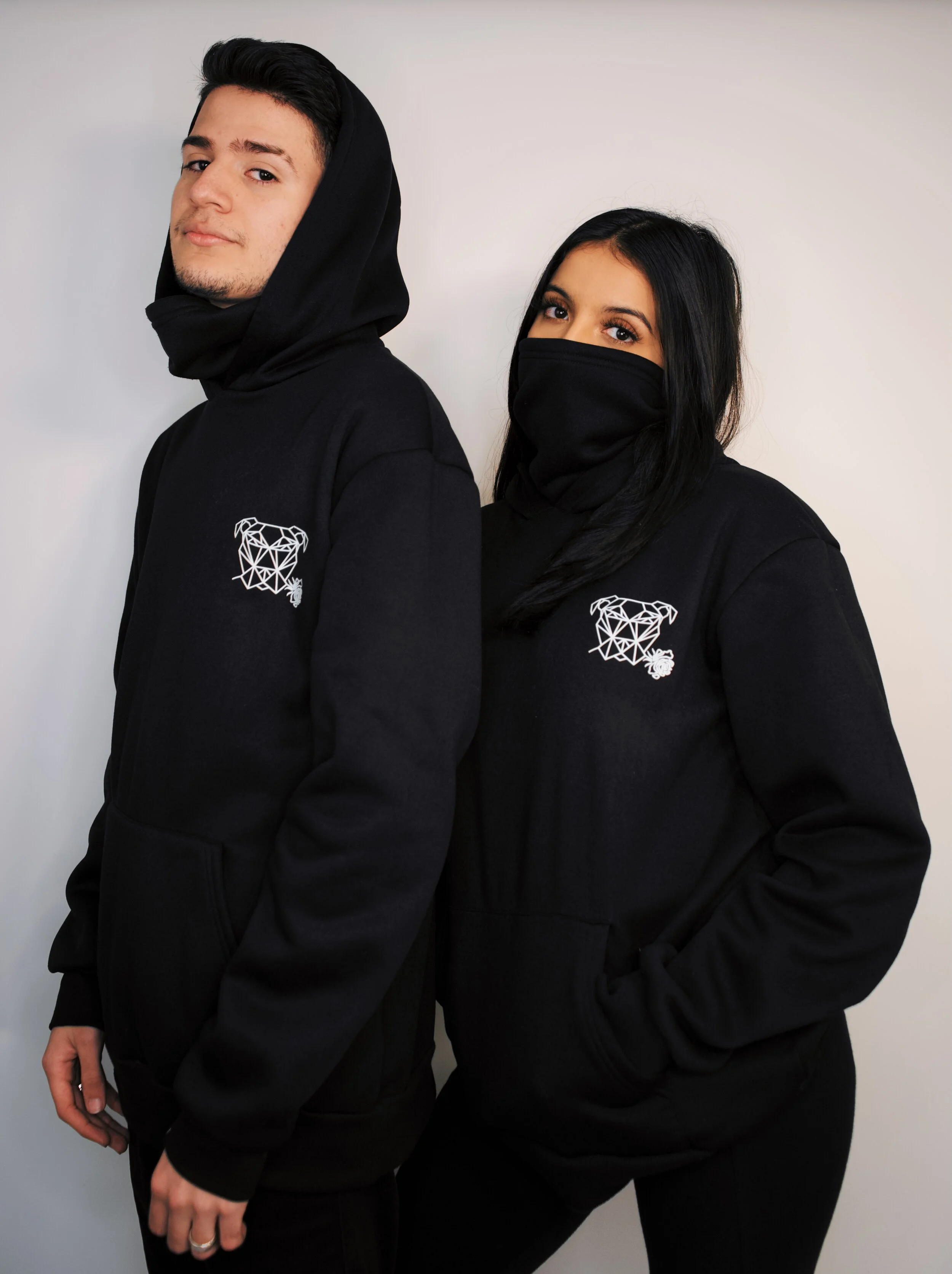 A young man and woman standing back to back, both wearing black hoodies with a geometric tiger face logo on the chest, and the woman also wearing a face covering. The background is plain white.