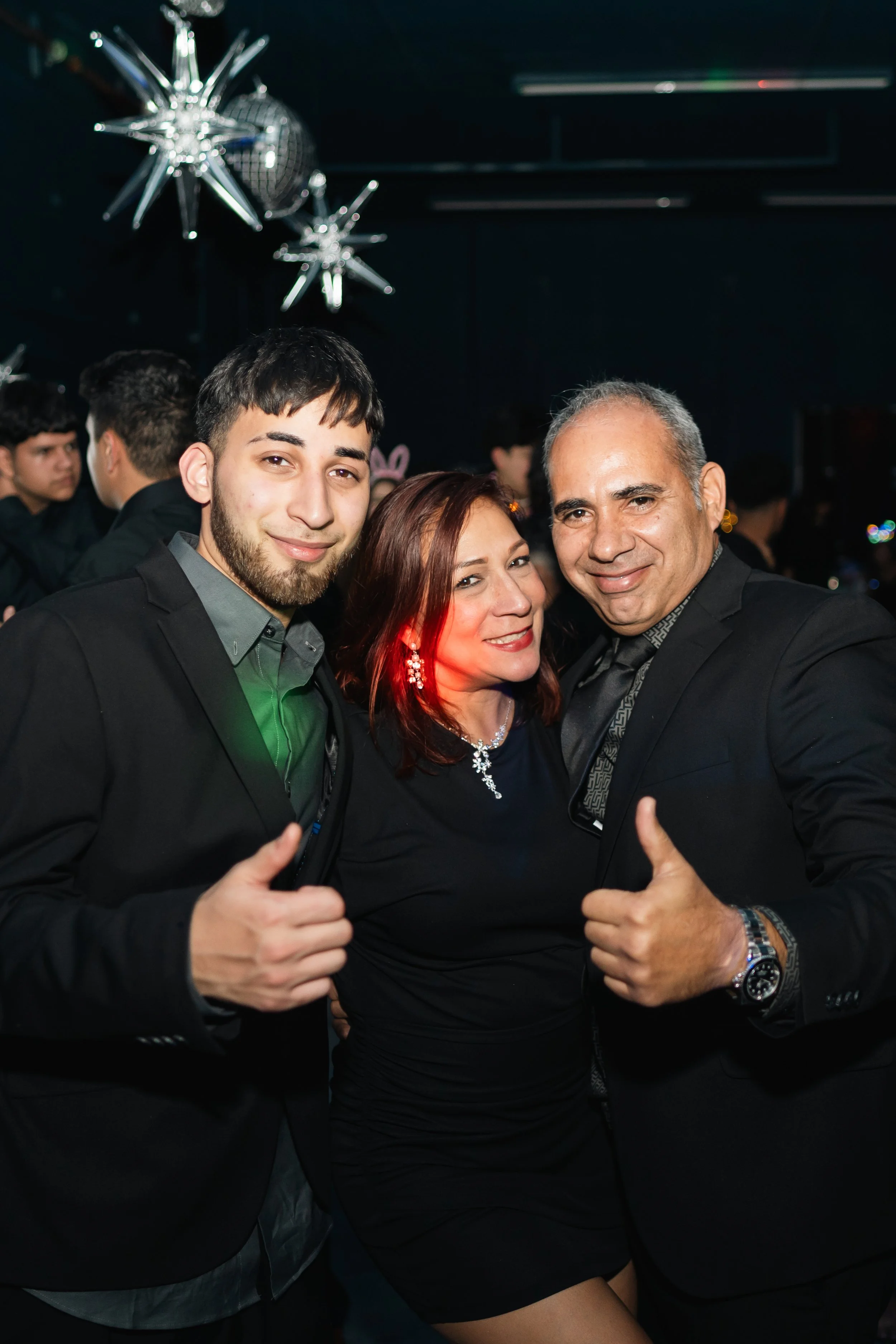 Three people smiling and giving thumbs up in a dark, festive setting with Christmas star decorations overhead.