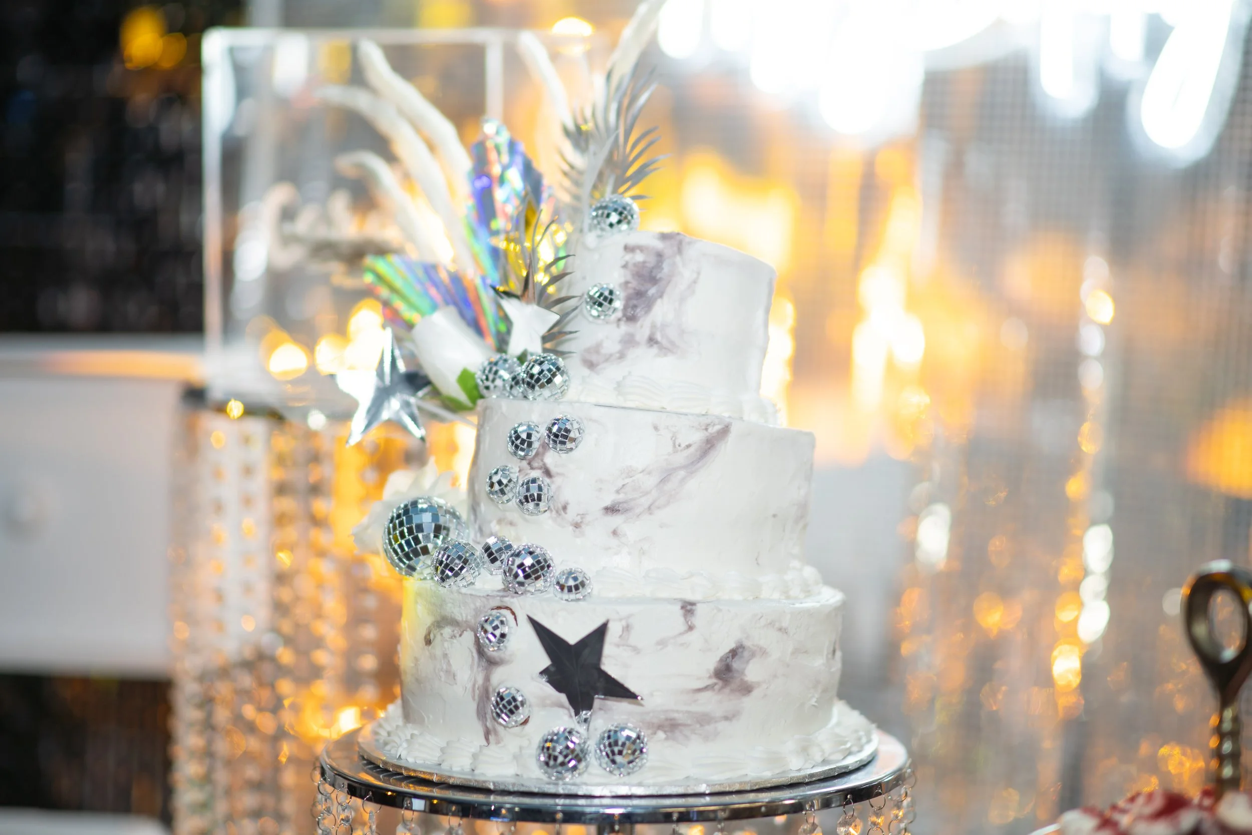 Three-tier white wedding cake decorated with disco ball ornaments, silver stars, and iridescent foliage, positioned on a reflective cake stand with a blurred background of warm lights and a diamond-like curtain.