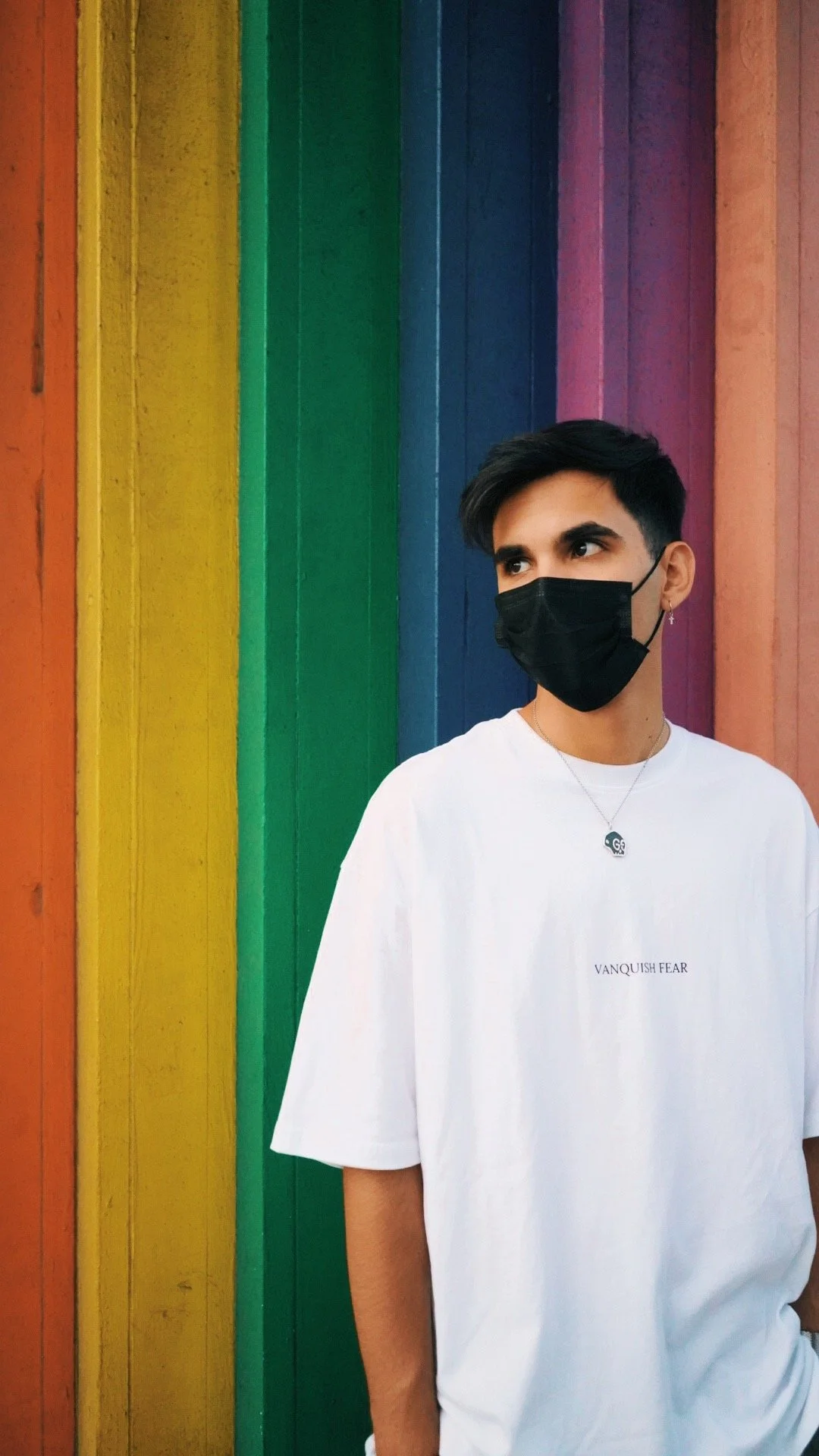 A young man wearing a black face mask, a white T-shirt with the words 'VANQUISH FEAR' on it, a necklace with a small pendant, and an earring stands in front of a colorful rainbow-striped wall.