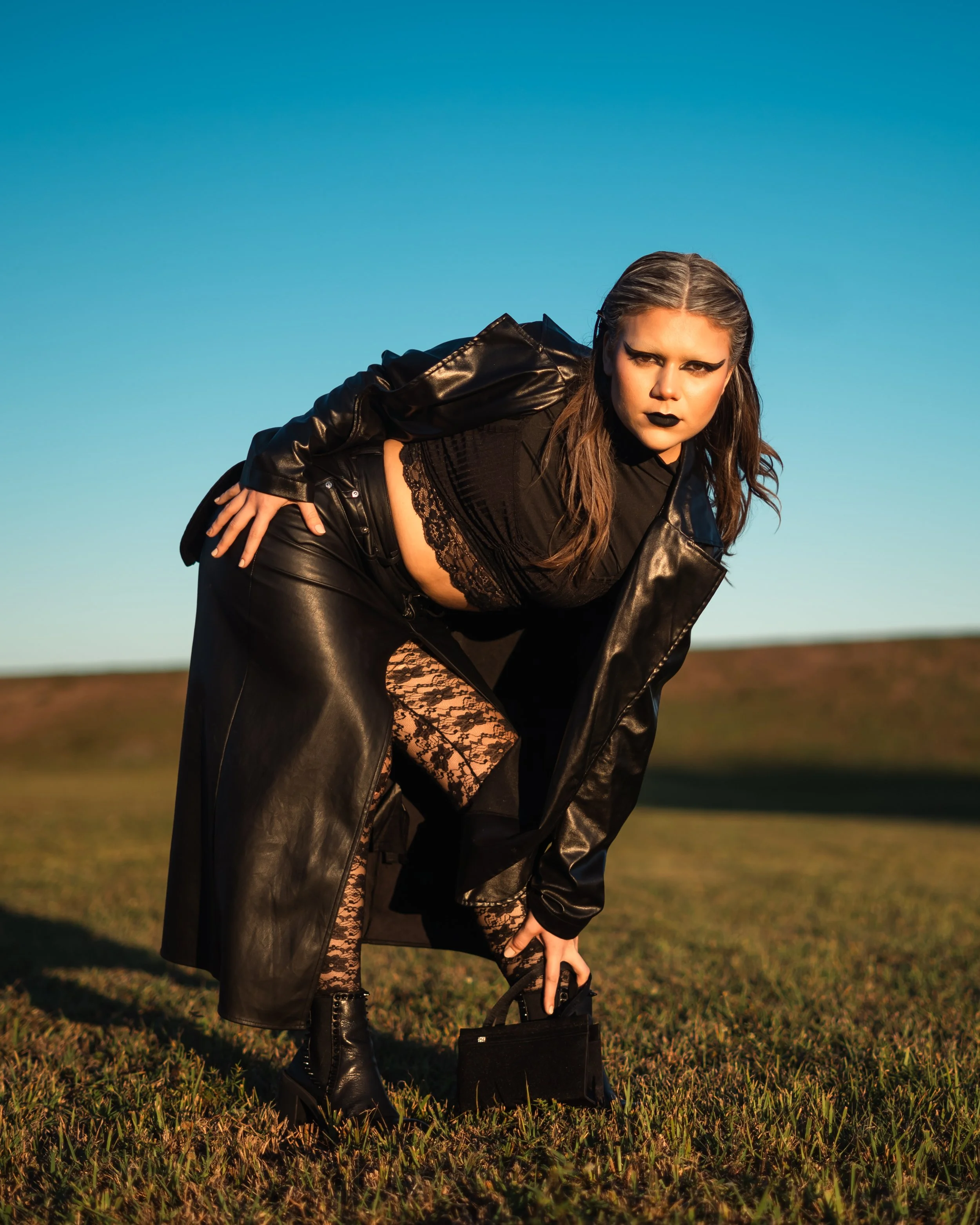 A woman with dark makeup, wearing a black leather coat, lace top, and lace pants, crouches on grass holding a small black handbag in an open outdoor setting with a clear blue sky.