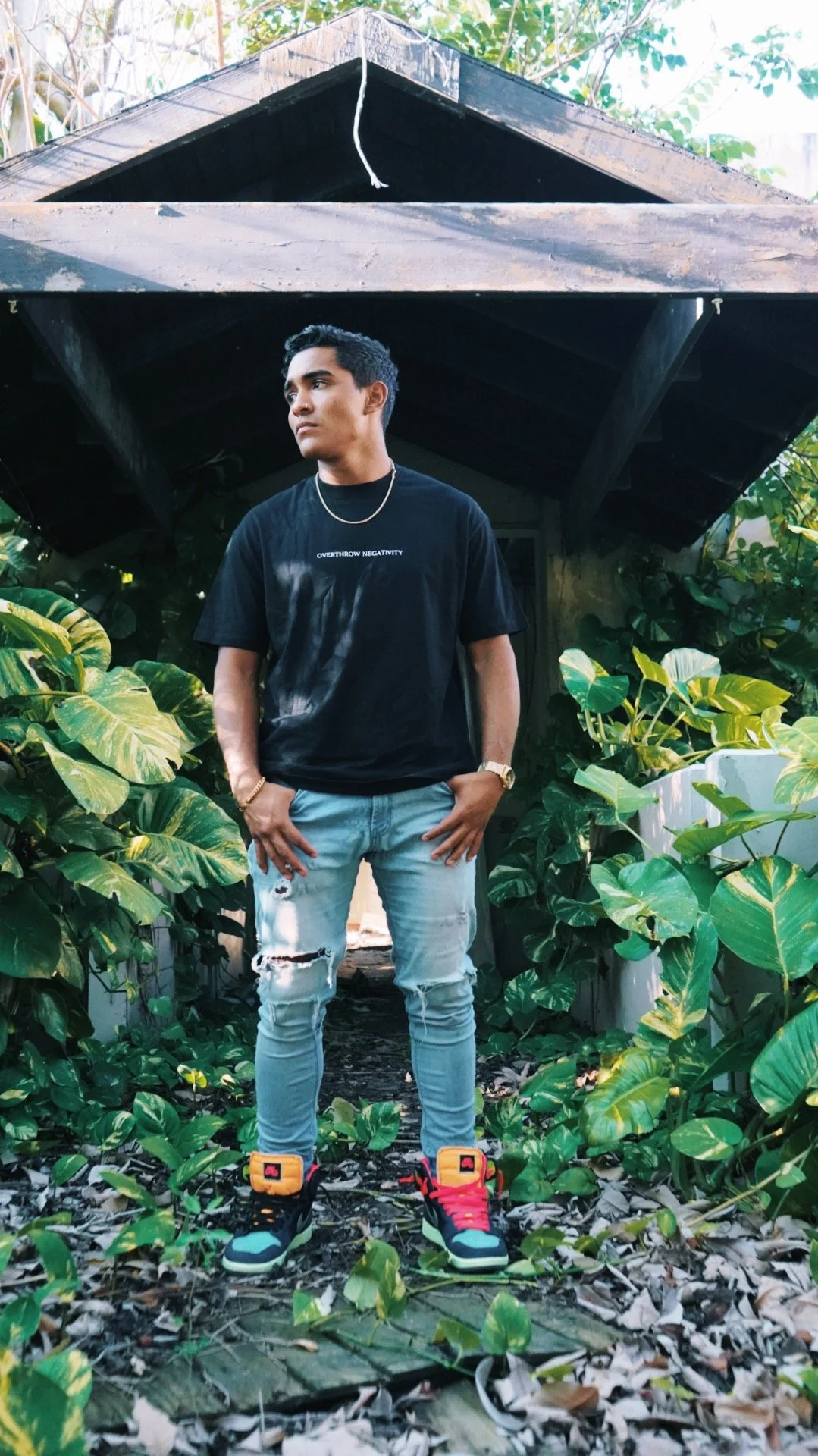 A young man stands in front of a small house or shed surrounded by green plants and leaves. He is wearing a black T-shirt, distressed light-wash jeans, and sneakers. His hands are in his pockets, and he has a serious expression.