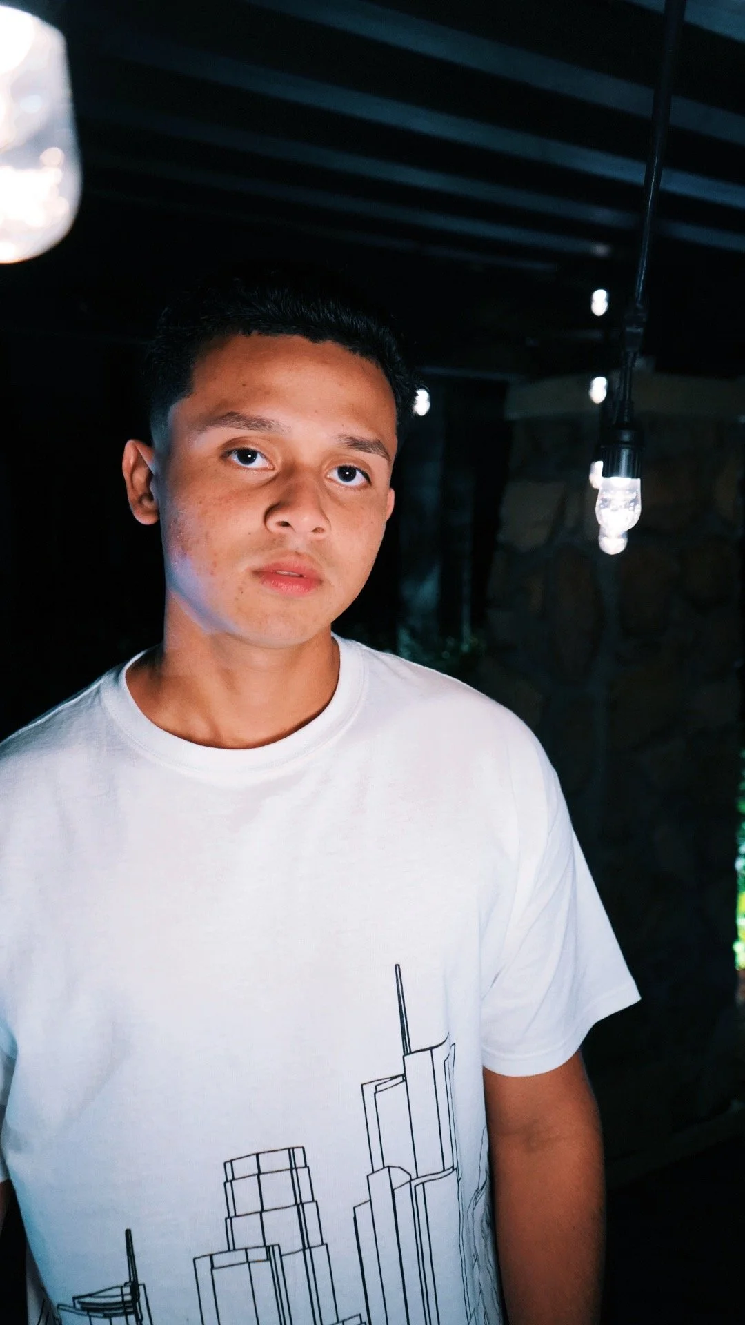 A young man with short dark hair and medium skin tone standing indoors in front of a dark background with string lights hanging from the ceiling, wearing a white T-shirt with a black line drawing of a city skyline.