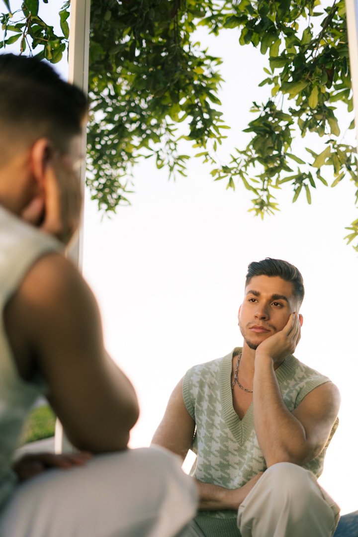 A man with styled dark hair and a sleeveless patterned sweater resting his face on his hand, appears to be listening thoughtfully to another person, with greenery overhead and bright sky in the background.