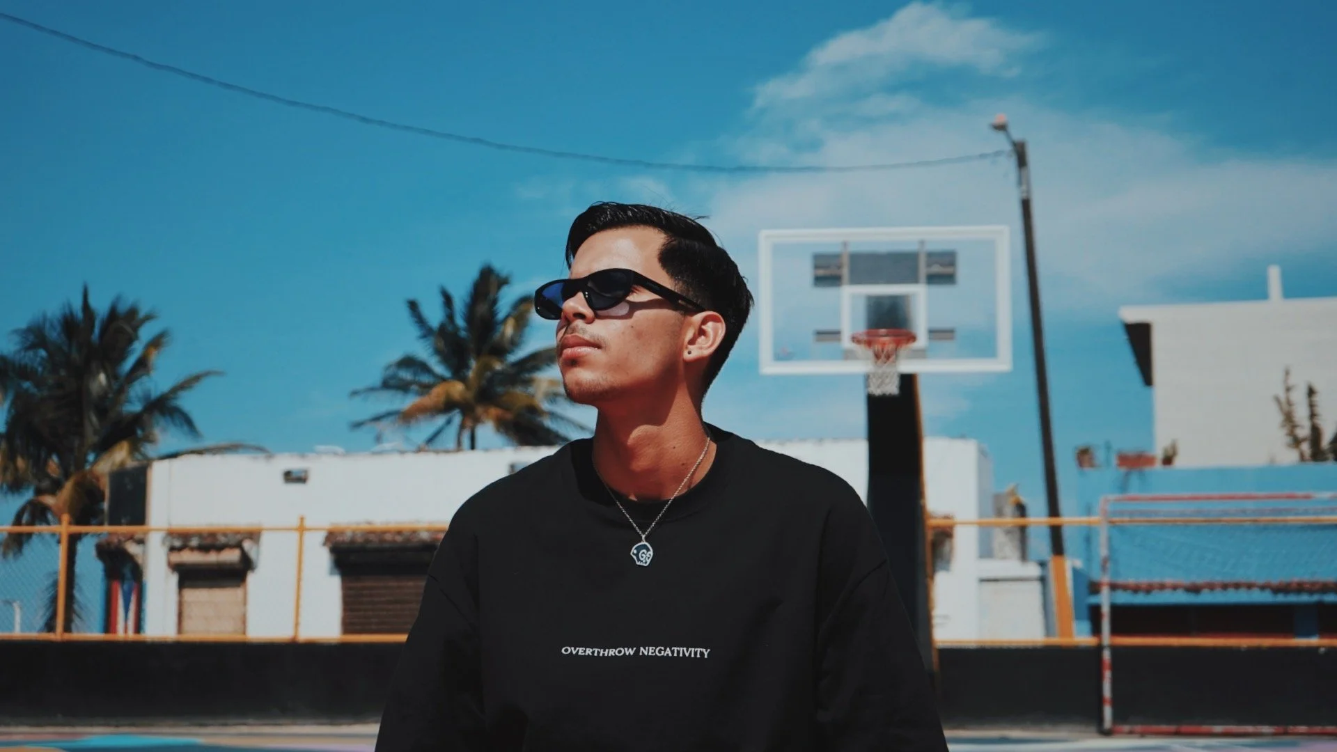 A young man with dark hair wearing sunglasses and a black shirt with white text, standing on an outdoor basketball court with a hoop, palm trees, and a blue sky in the background.