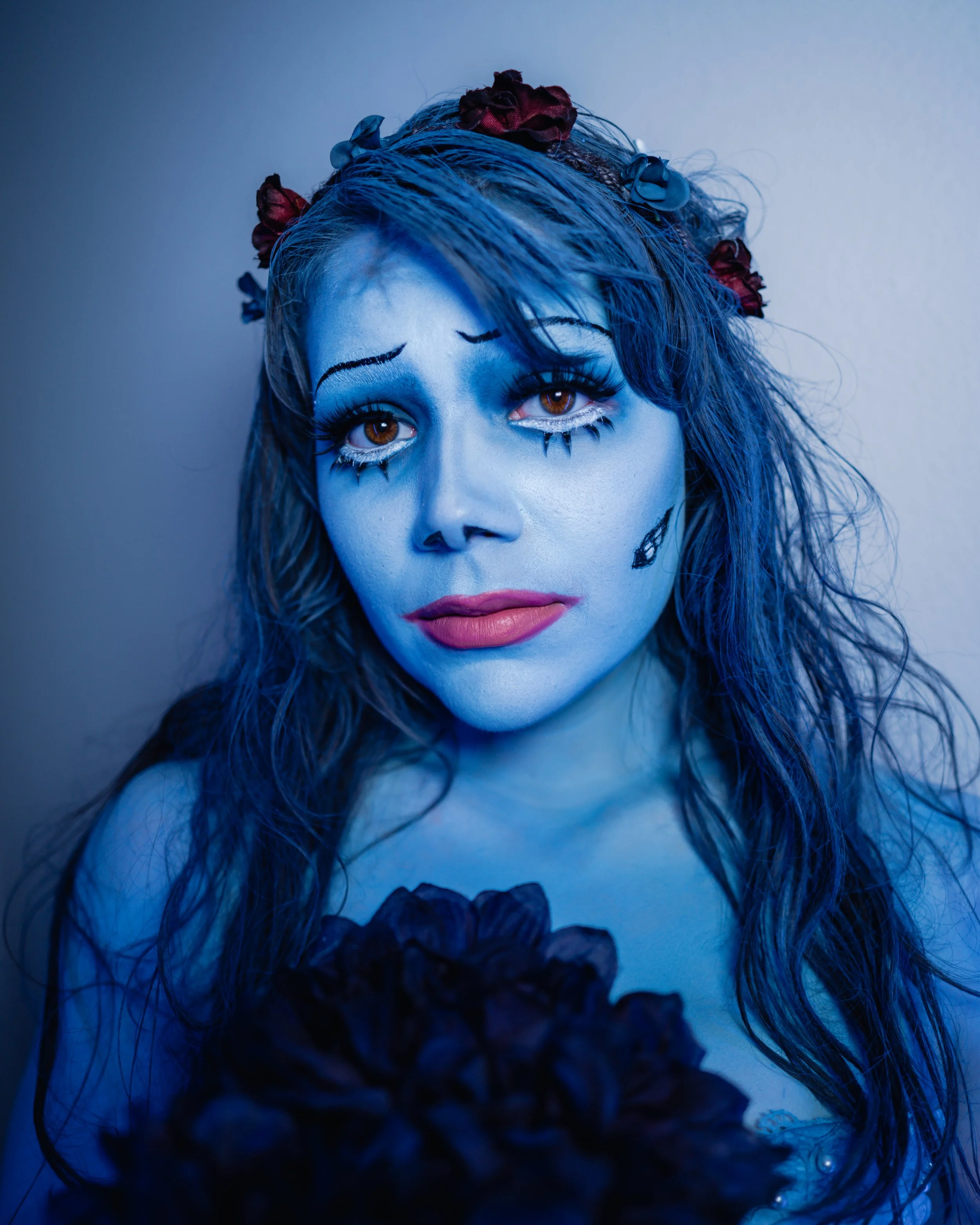 Woman with blue face paint and theatrical makeup, wearing a flower crown and dark clothing, against a plain background.