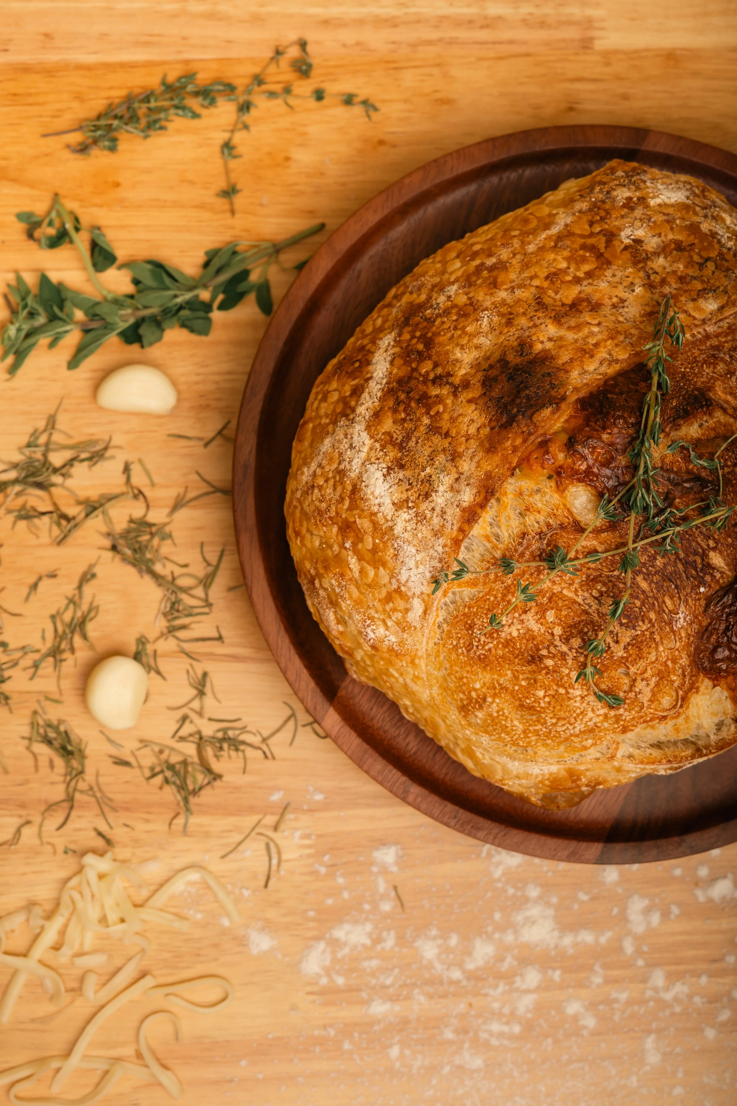 A loaf of bread on a wooden plate surrounded by garlic cloves, fresh herbs, and uncooked pasta on a wooden surface.