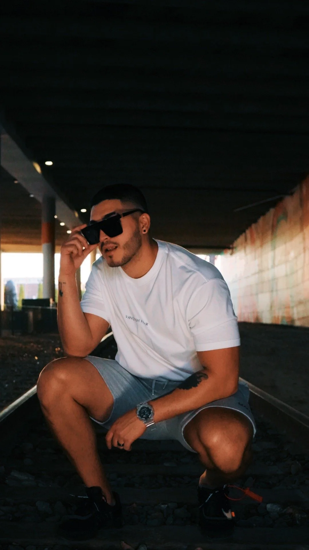 A young man with dark hair and a beard sitting on train tracks under a bridge, wearing sunglasses, a white T-shirt, gray shorts, black sneakers, a watch, and a ring, squatting with one knee on the ground.