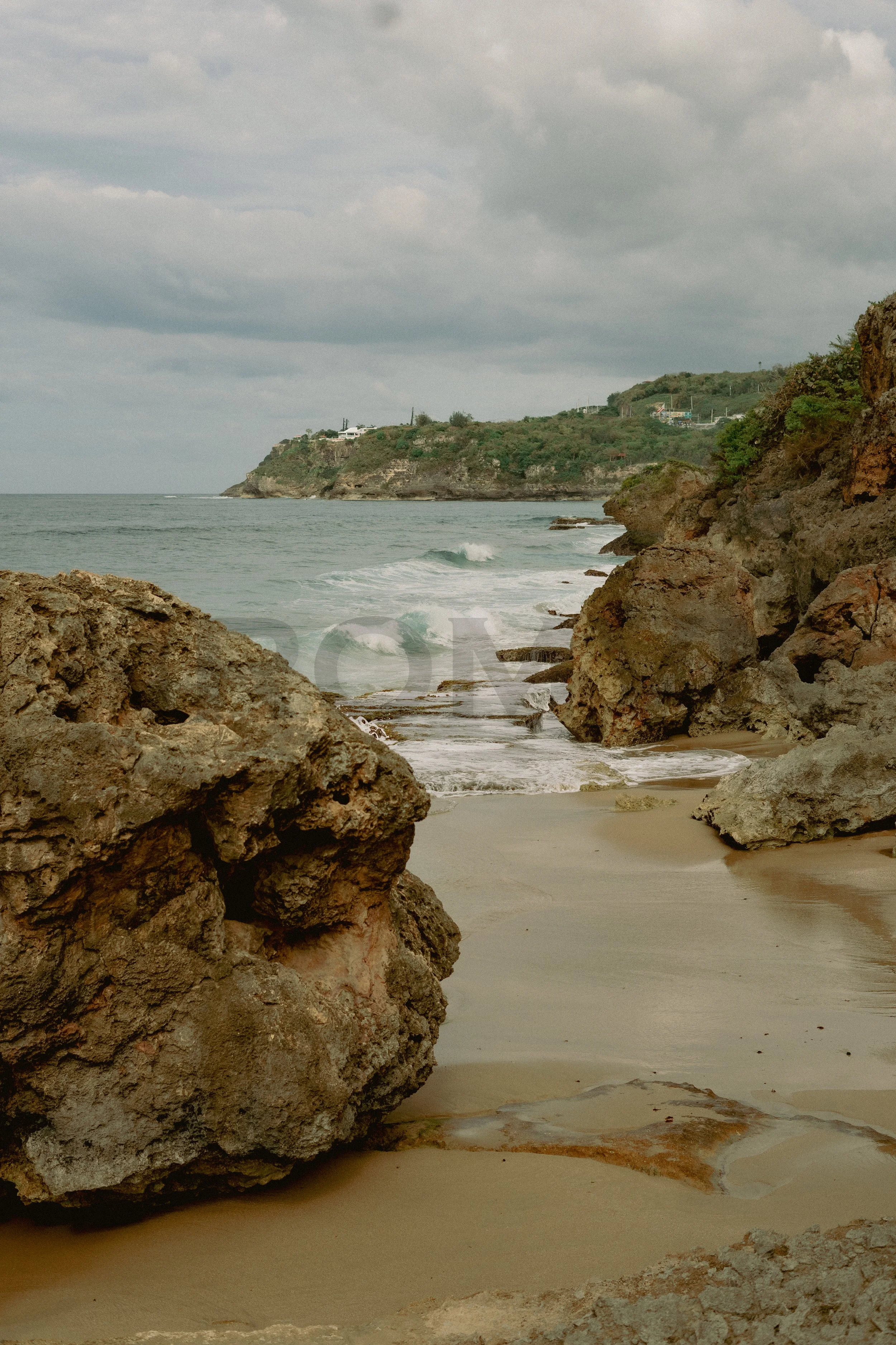 A rocky beach with large boulders and waves hitting the shore, cliffs in the background under a cloudy sky.