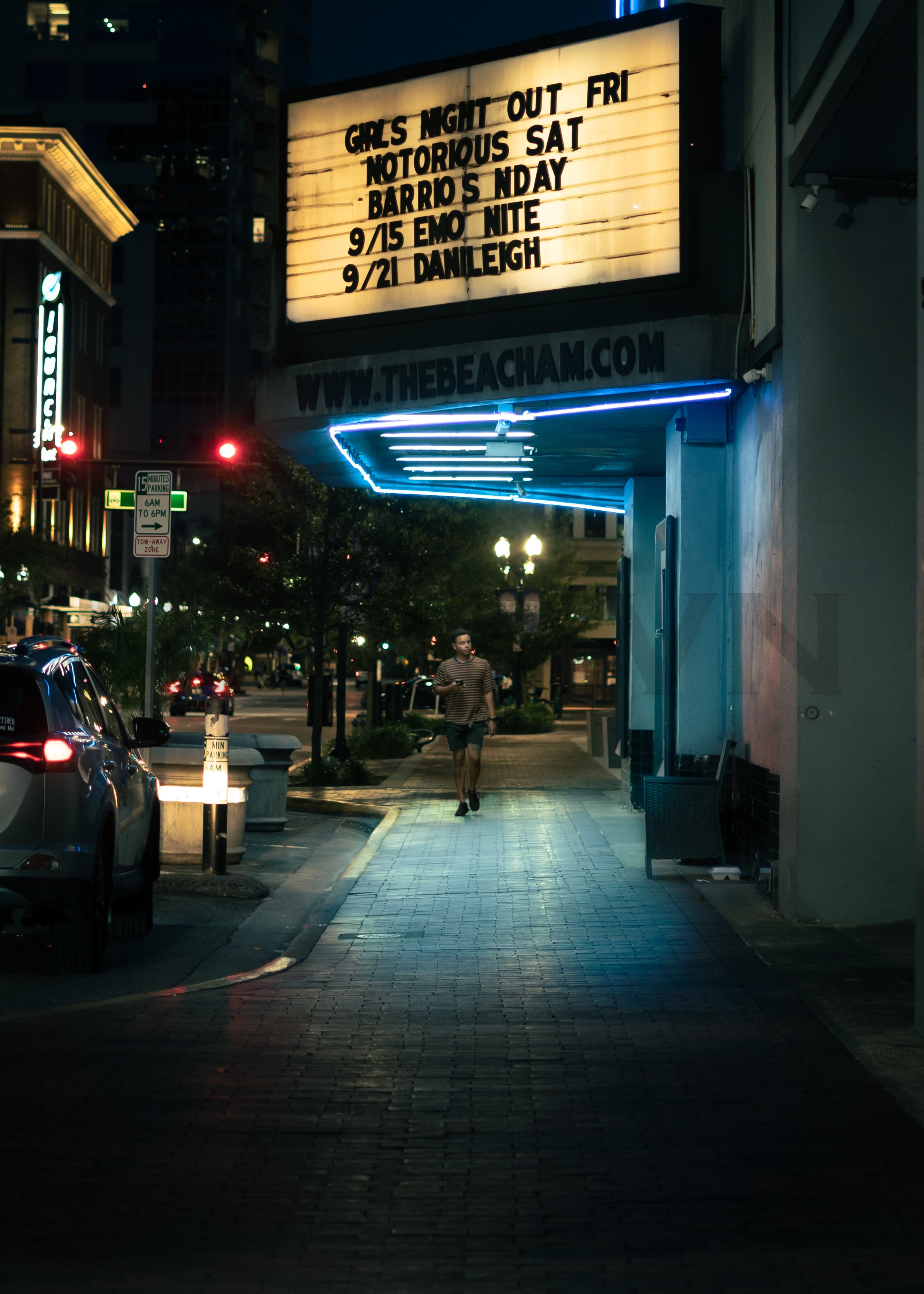 A nighttime street scene with a man walking on the sidewalk. There is a marquee sign with event listings and a website URL, illuminated by neon lights, and cars parked along the street.