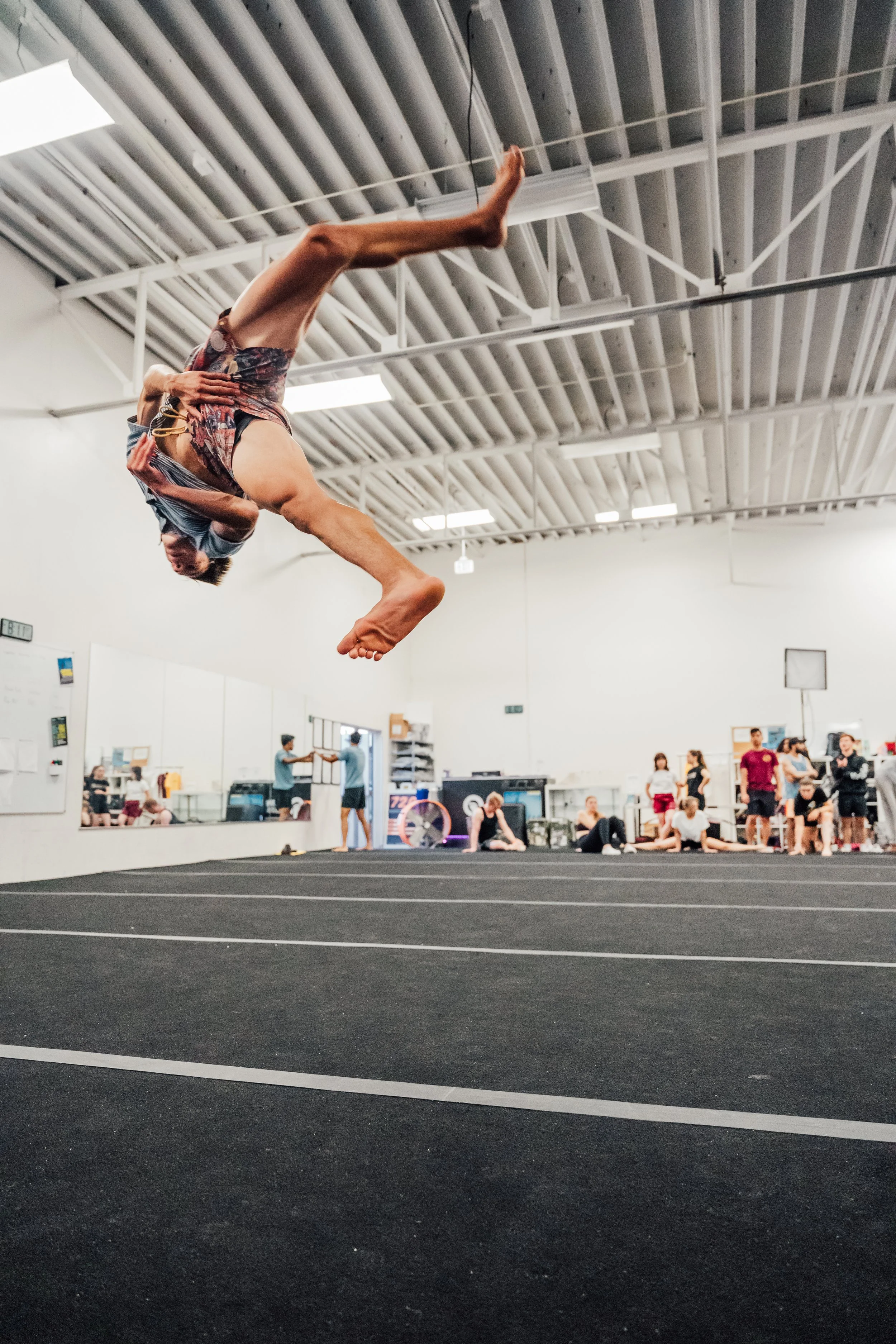 A person performing a backflip inside a gym with spectators and coaches watching.