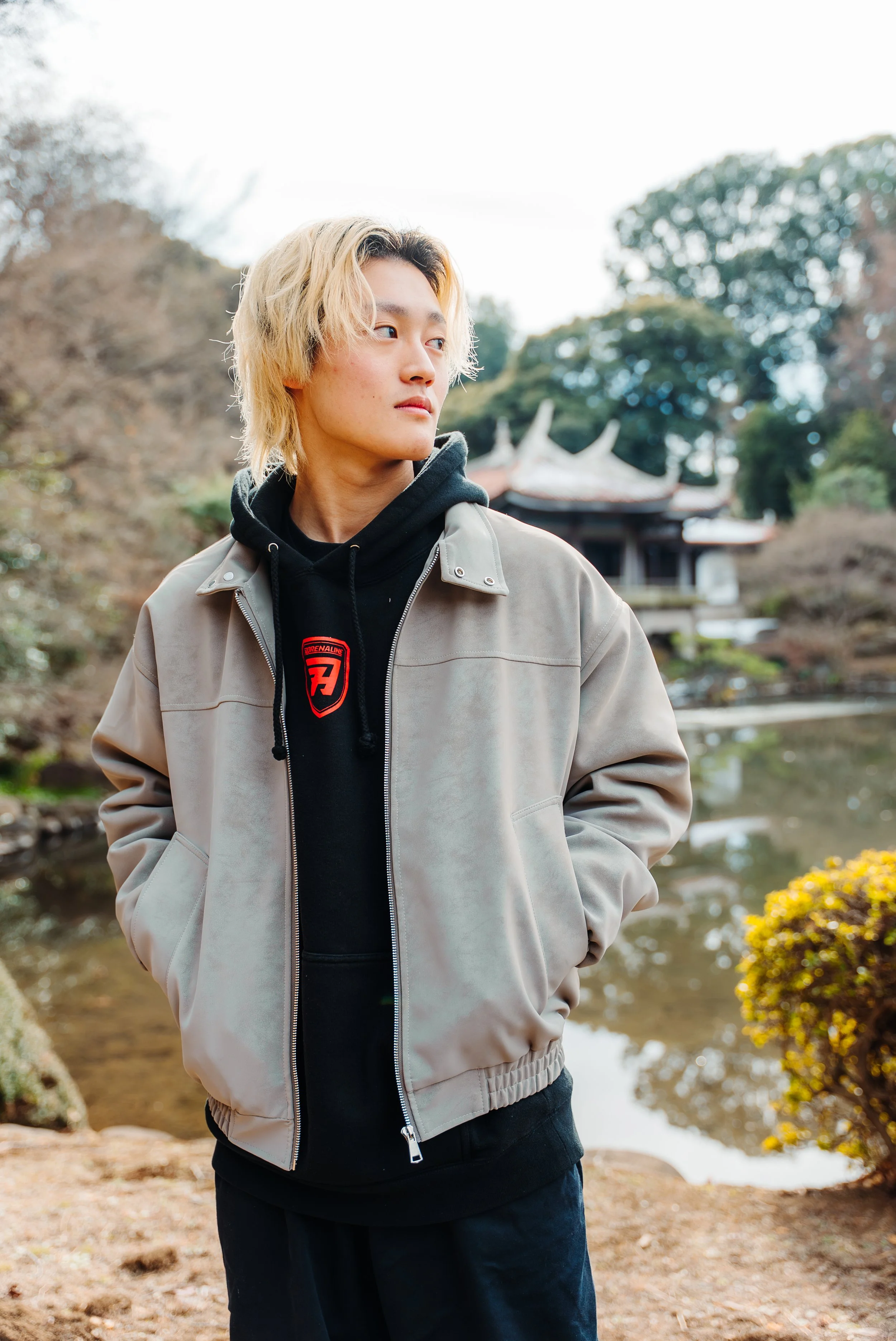 A young man with blond hair stands outdoors near a pond with trees and a traditional Asian pavilion in the background, wearing a beige jacket over a black hoodie.