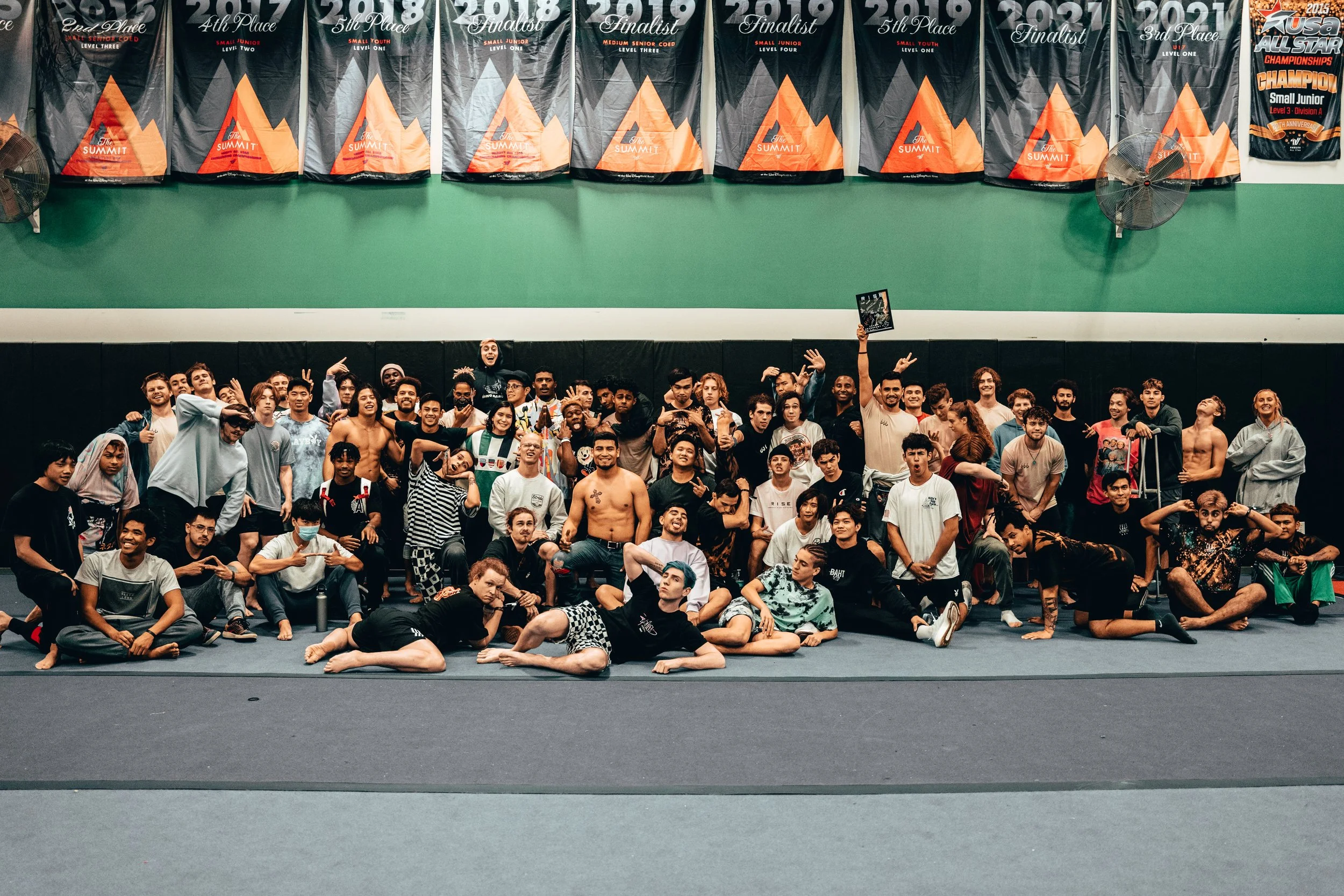 A large group of diverse people, including men and women, posing together in an indoor sports facility with banners hanging on the wall that display various sports achievements and rankings.