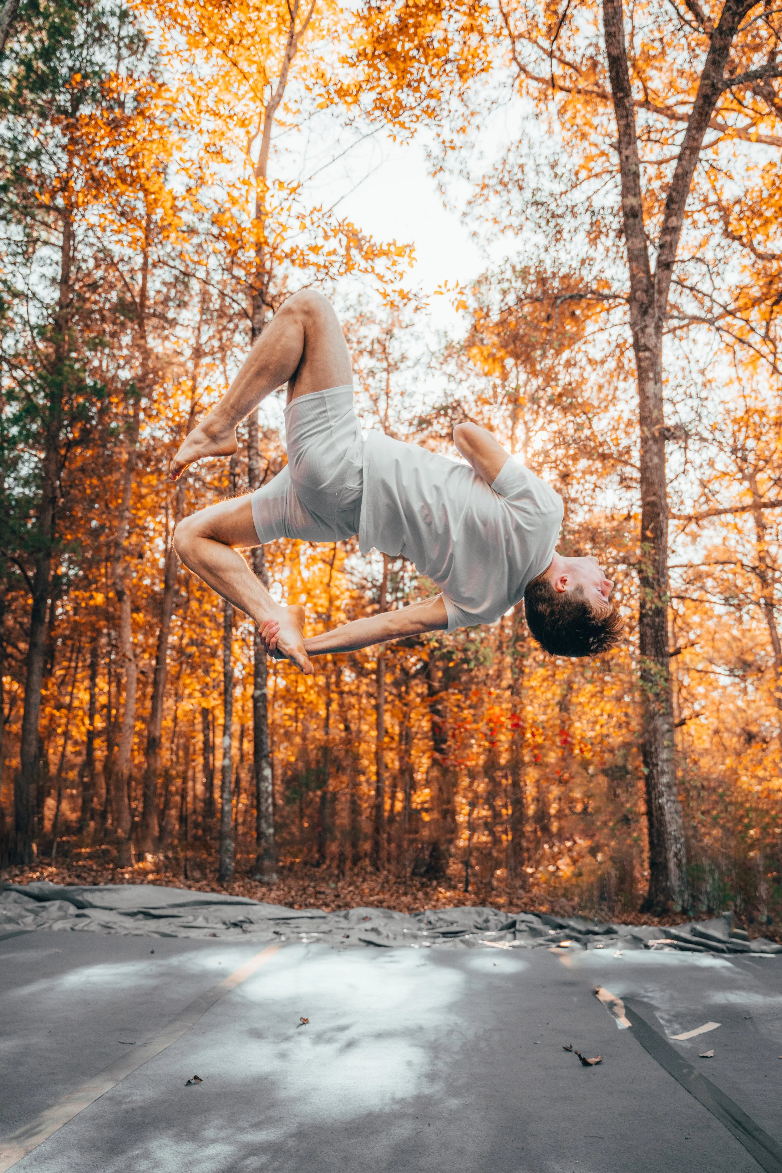 A man in a white t-shirt and shorts performing a flip on a trampoline in a forest with autumn-colored leaves.