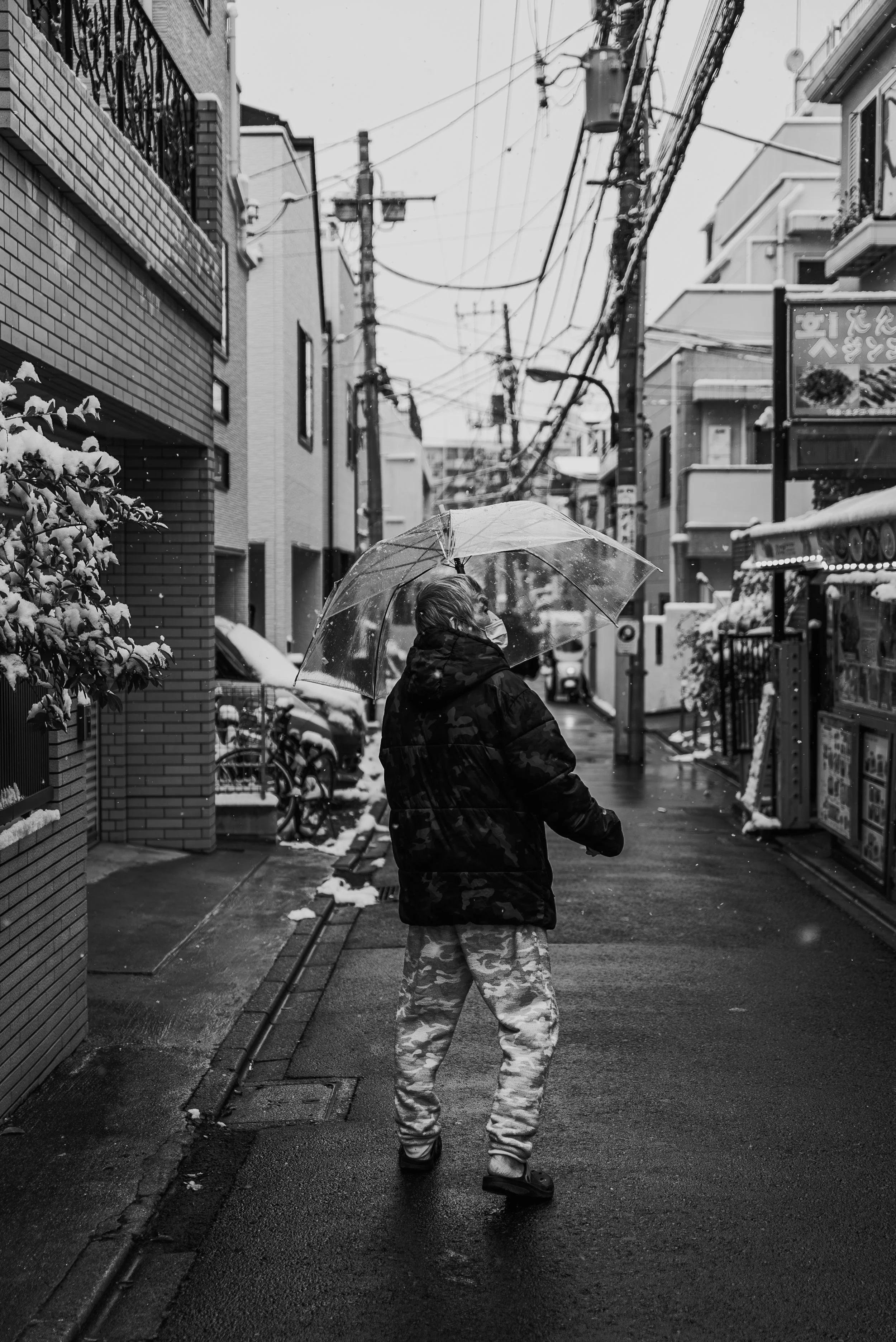 A person walking on a rainy street holding a transparent umbrella. They are wearing a black jacket and camouflage pants. Snow is visible on the ground and on nearby objects. Buildings and overhead utility wires are in the background, with some signs 