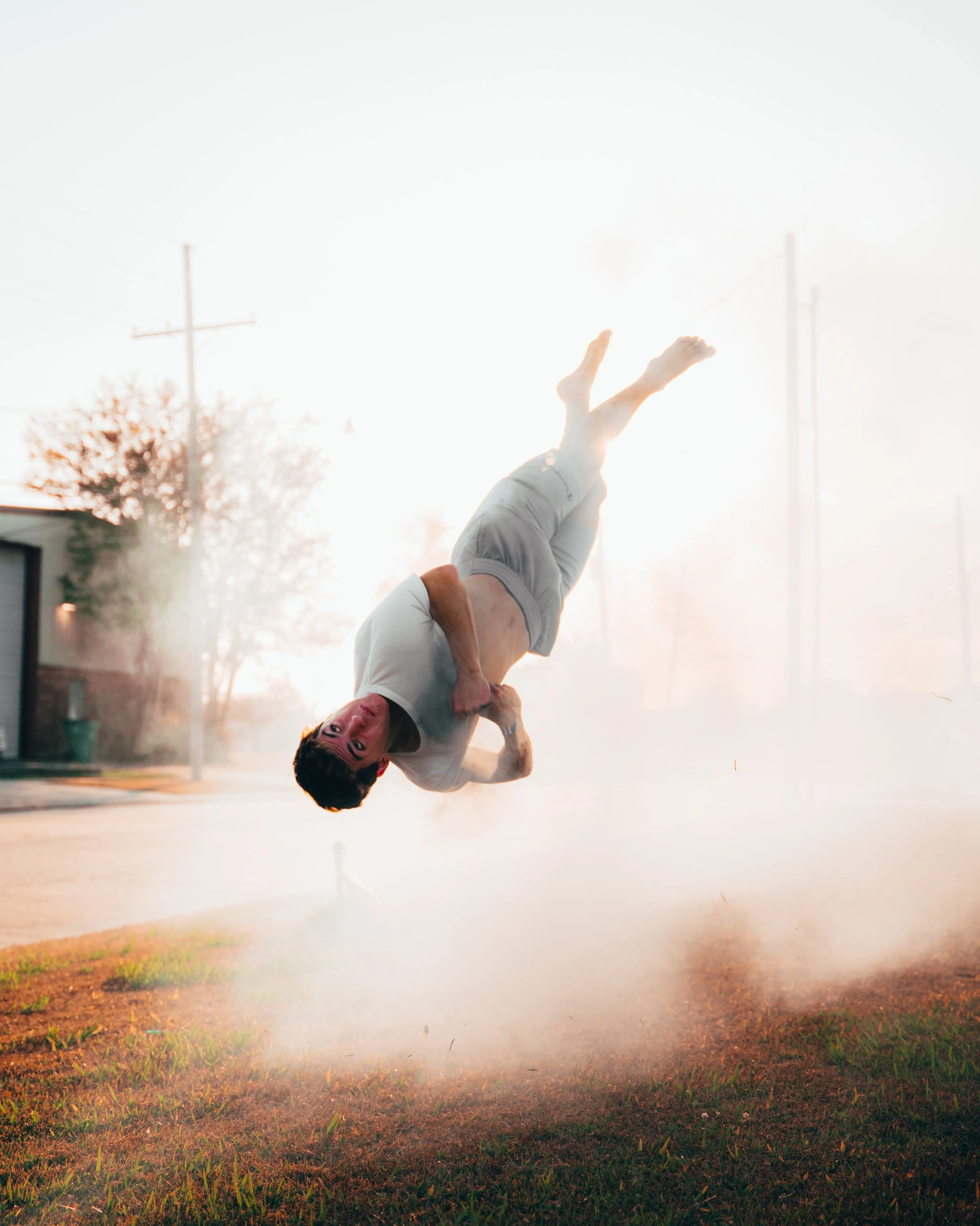 A person falling or flipping during a stunt on a street, with dust or smoke surrounding them in a bright outdoor setting at sunrise or sunset.