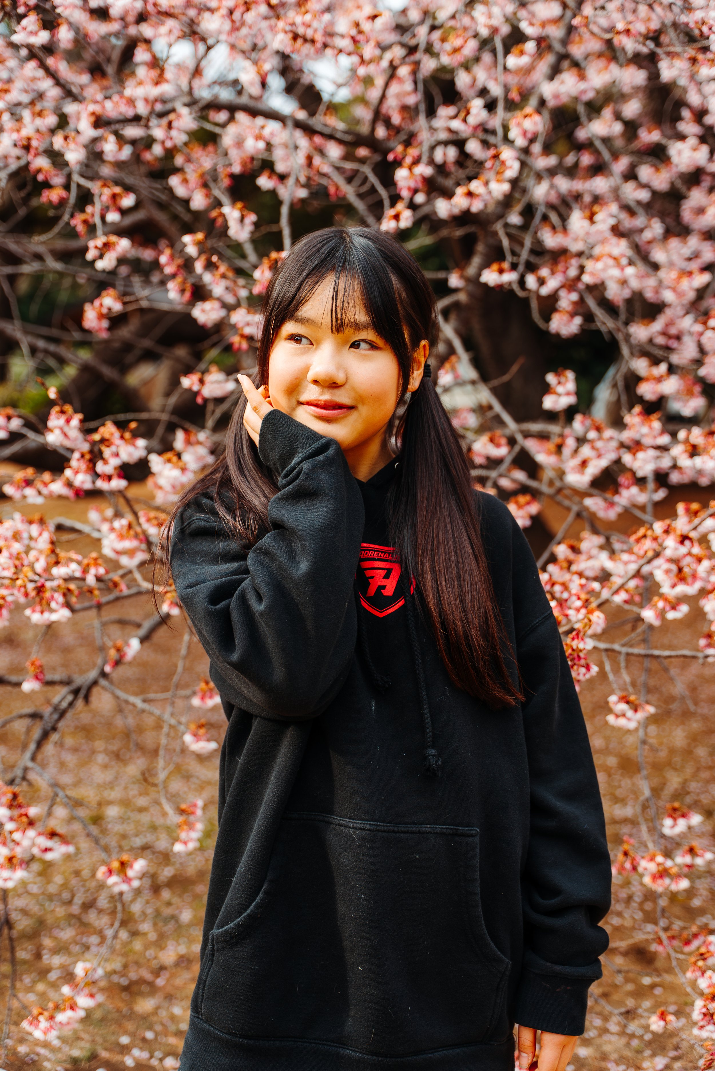 A young woman with long dark hair and a black hoodie standing in front of a blossoming cherry tree.