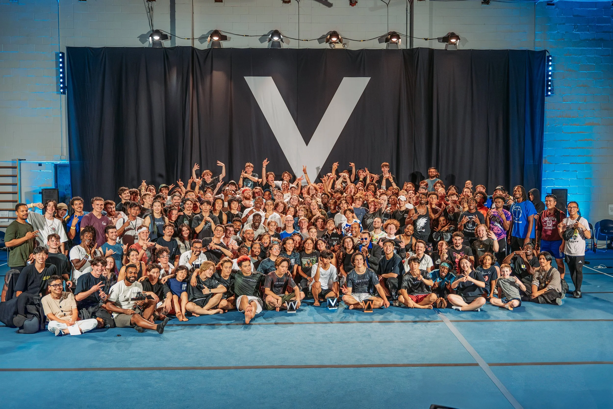 A large group of diverse people posing together in front of a black curtain with a large white 'V' on it, inside a gym or event space with blue flooring and industrial lighting.
