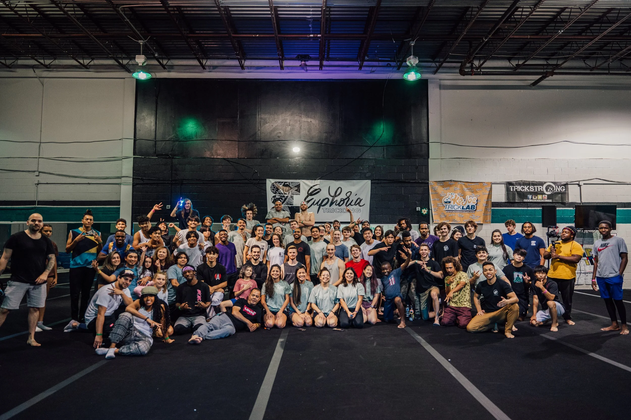 Large group of people in a gymnasium, many wearing casual clothes or athletic wear, posing for a group photo with some making peace signs or other gestures, with banners and signs in the background related to tricking and performing arts.