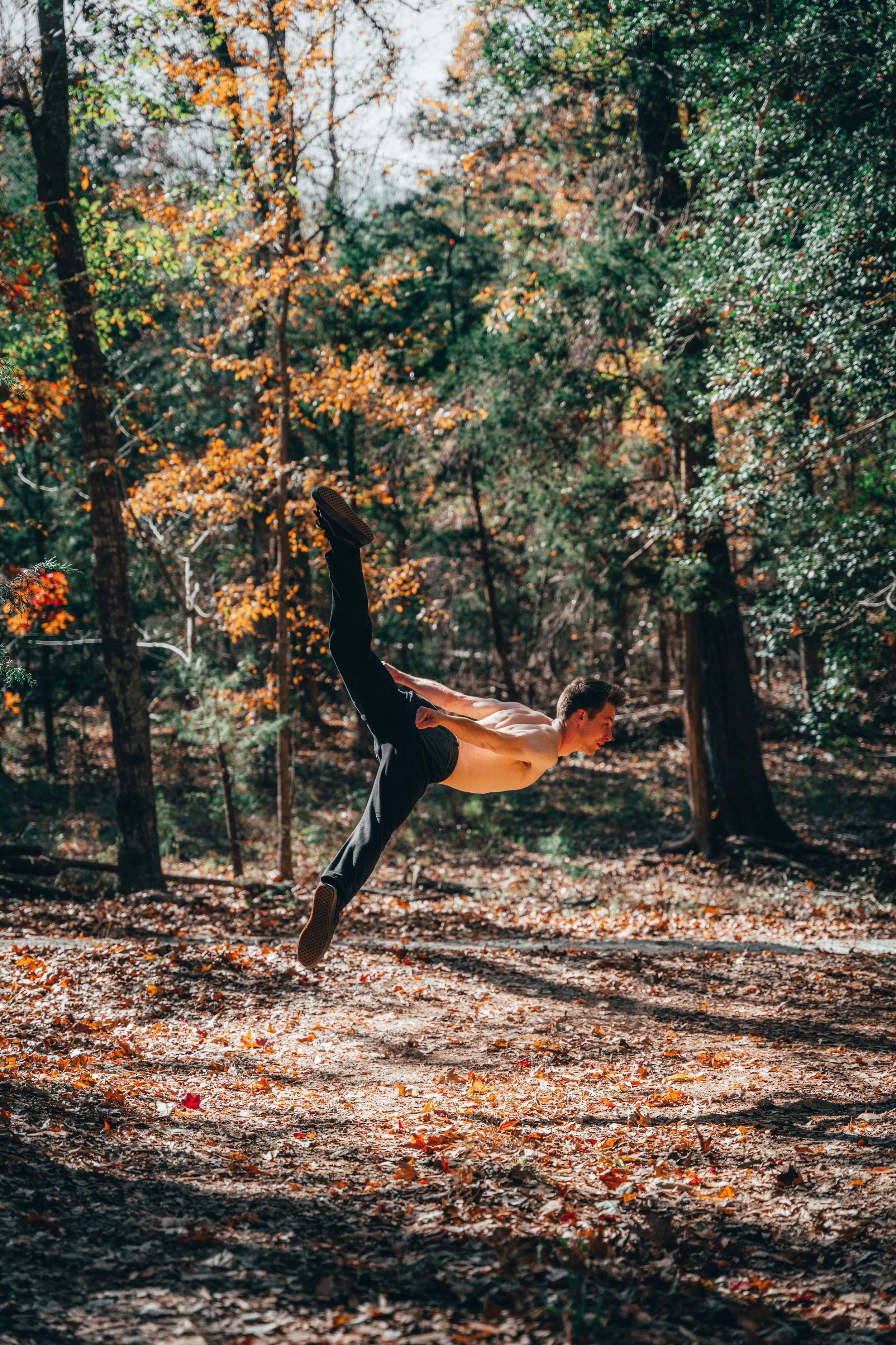 A shirtless man in black pants performing a back handspring in a forest with autumn foliage.